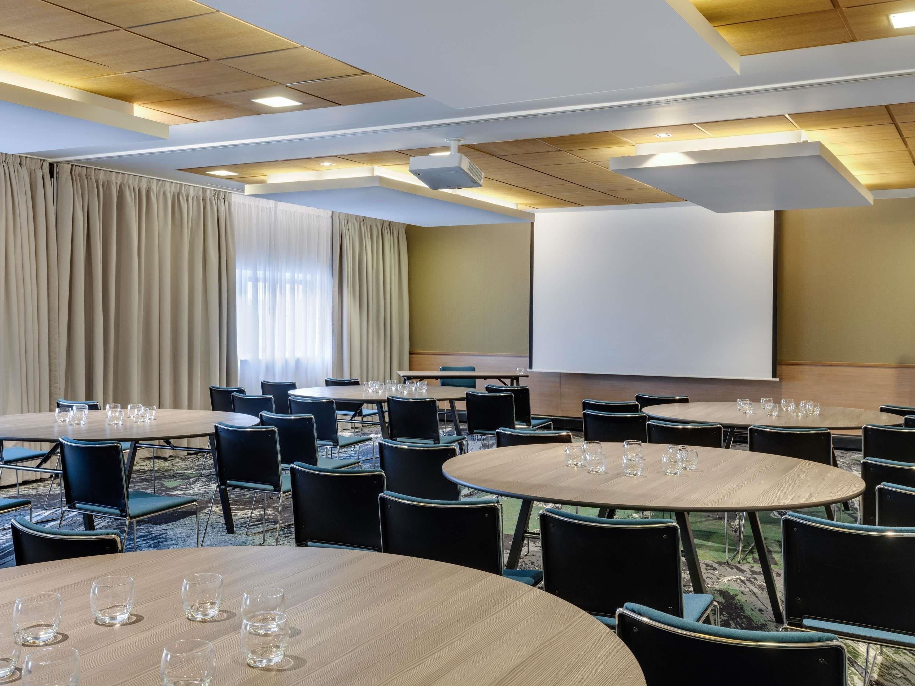 A conference room with multiple tables and chairs, a large screen, and a view of the outdoors.