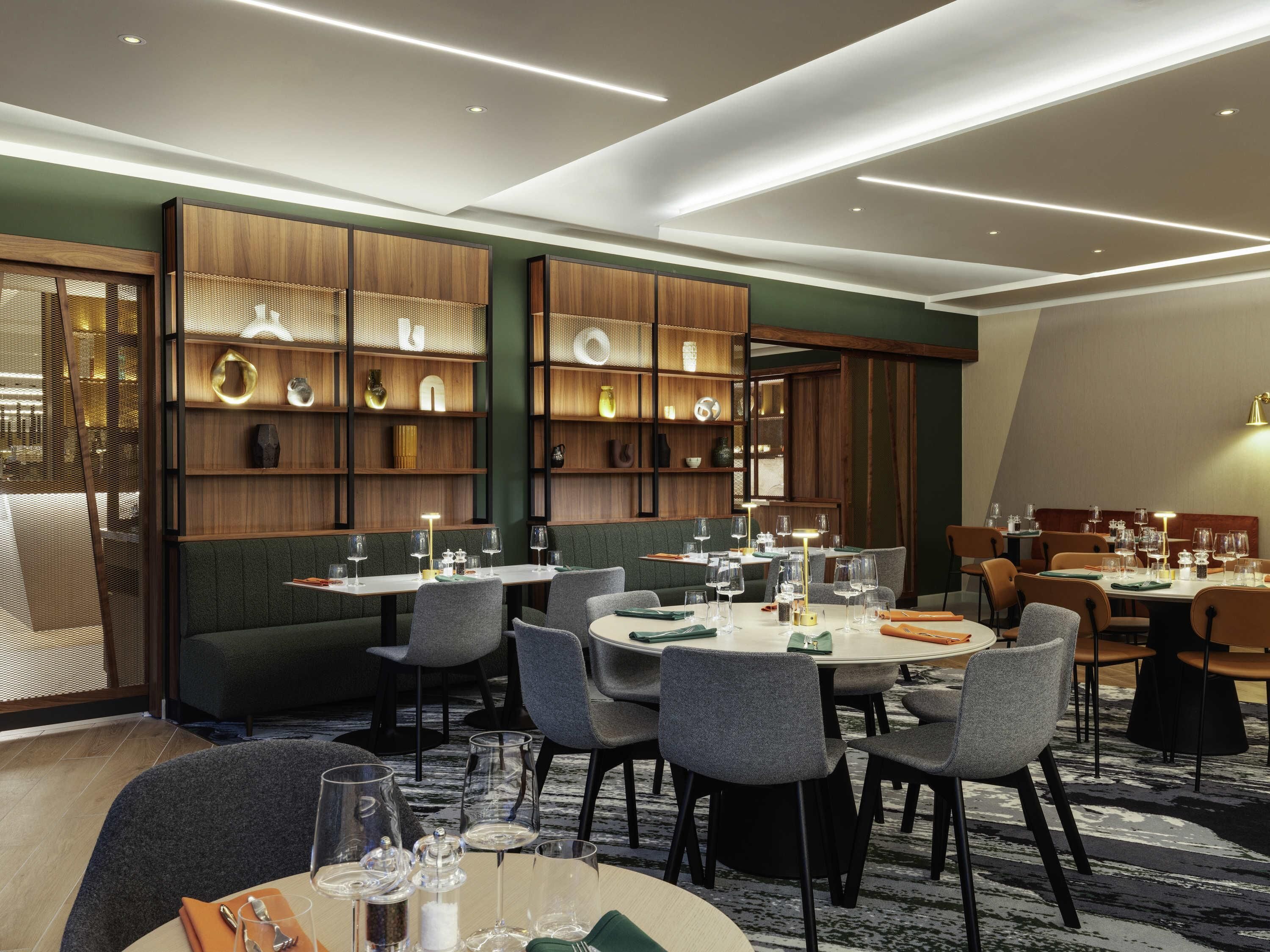 A modern dining area with round tables, grey upholstered chairs, and wooden shelving with decorative items.