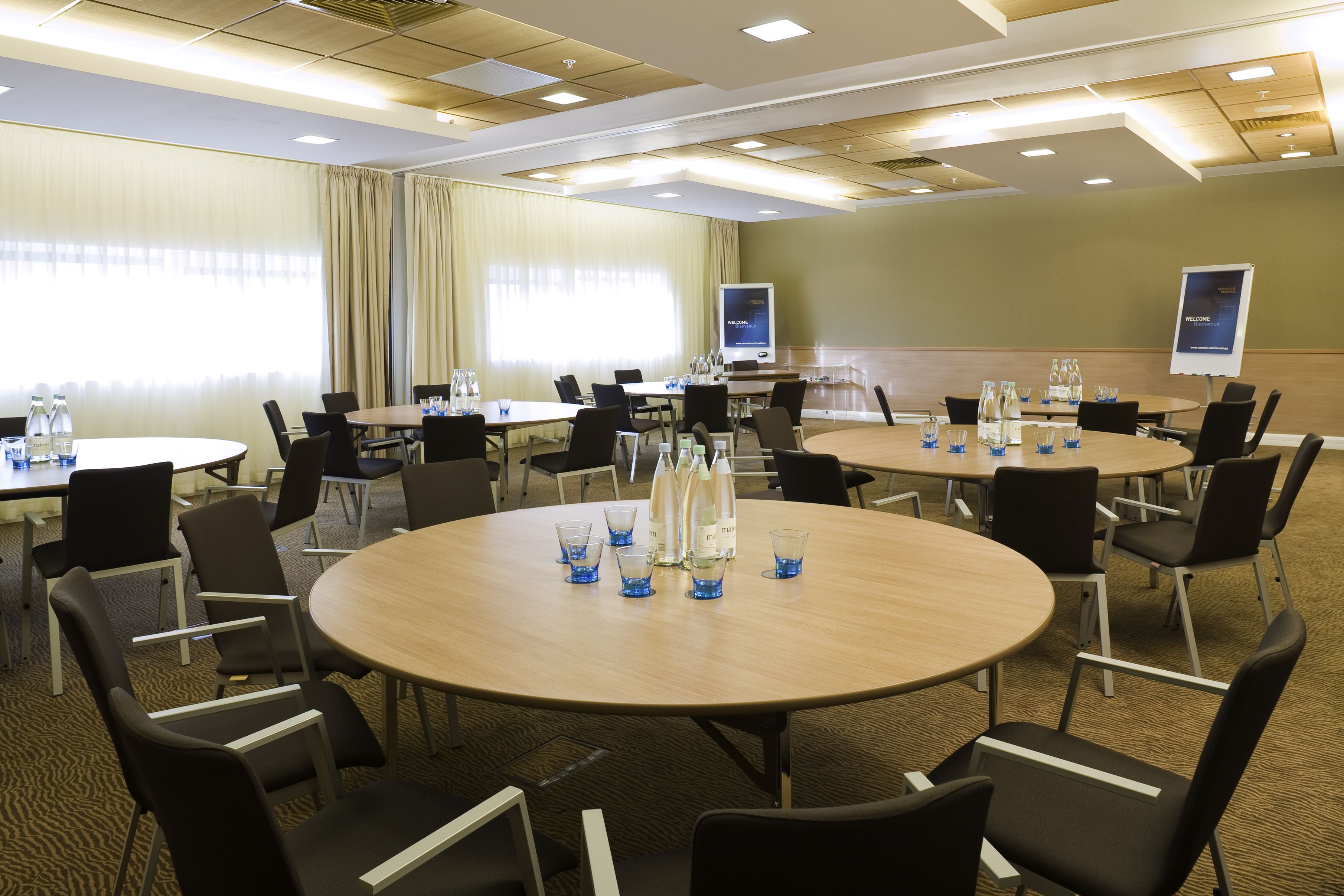 A conference room with multiple tables and chairs, equipped with water bottles and glasses, and a screen displaying a welcome message.