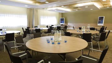 A conference room with multiple tables and chairs, equipped with water bottles and glasses, and a screen displaying a welcome message.