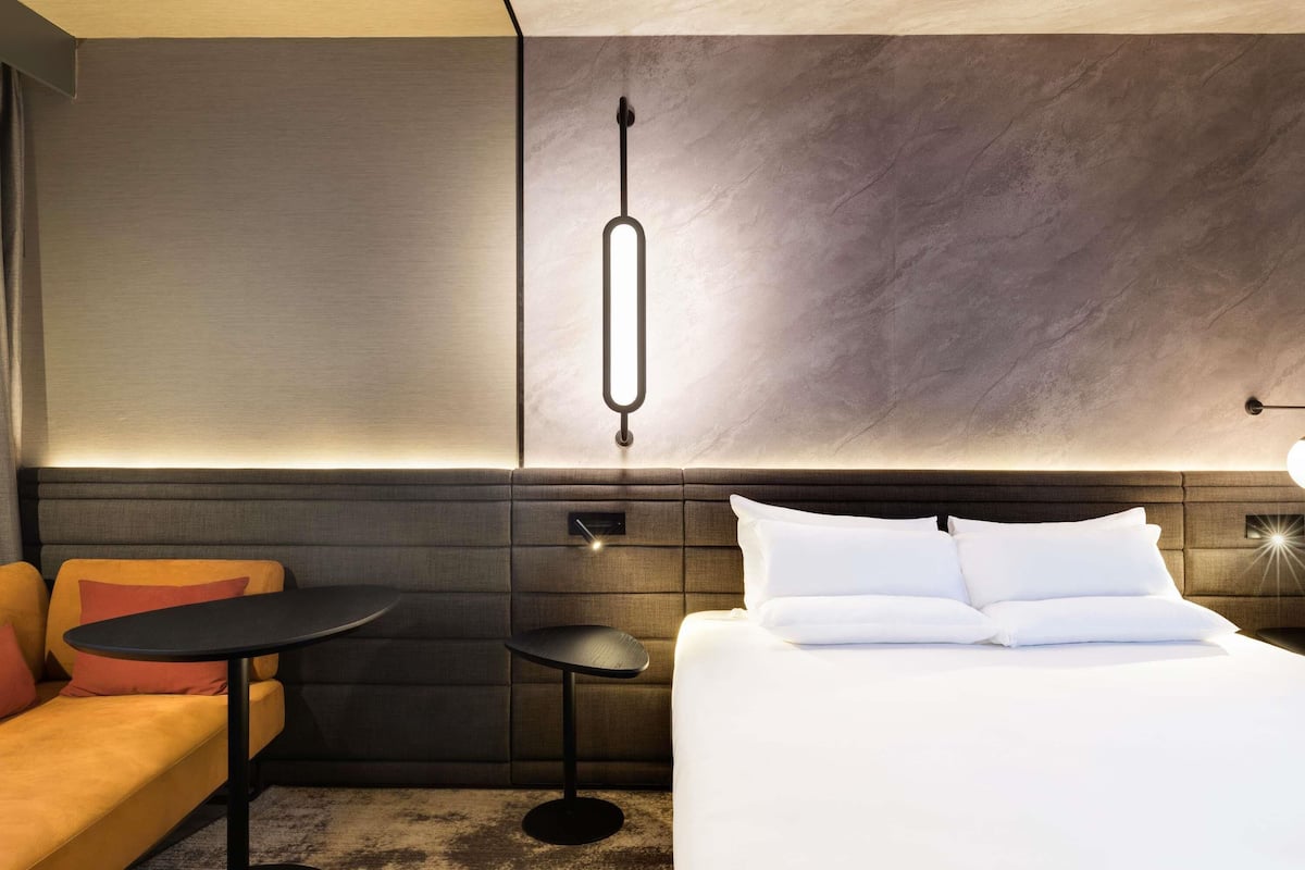 A modern hotel room with a bed, a small round table, a sofa, and a hanging light fixture.
