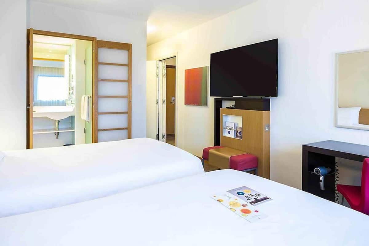A hotel room with a bed, a TV mounted on the wall, a desk with a chair, and a small seating area.