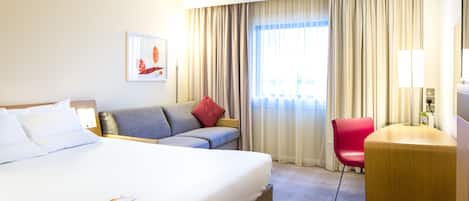 A hotel room with a large bed, a sofa, a red chair, and a desk with a lamp.