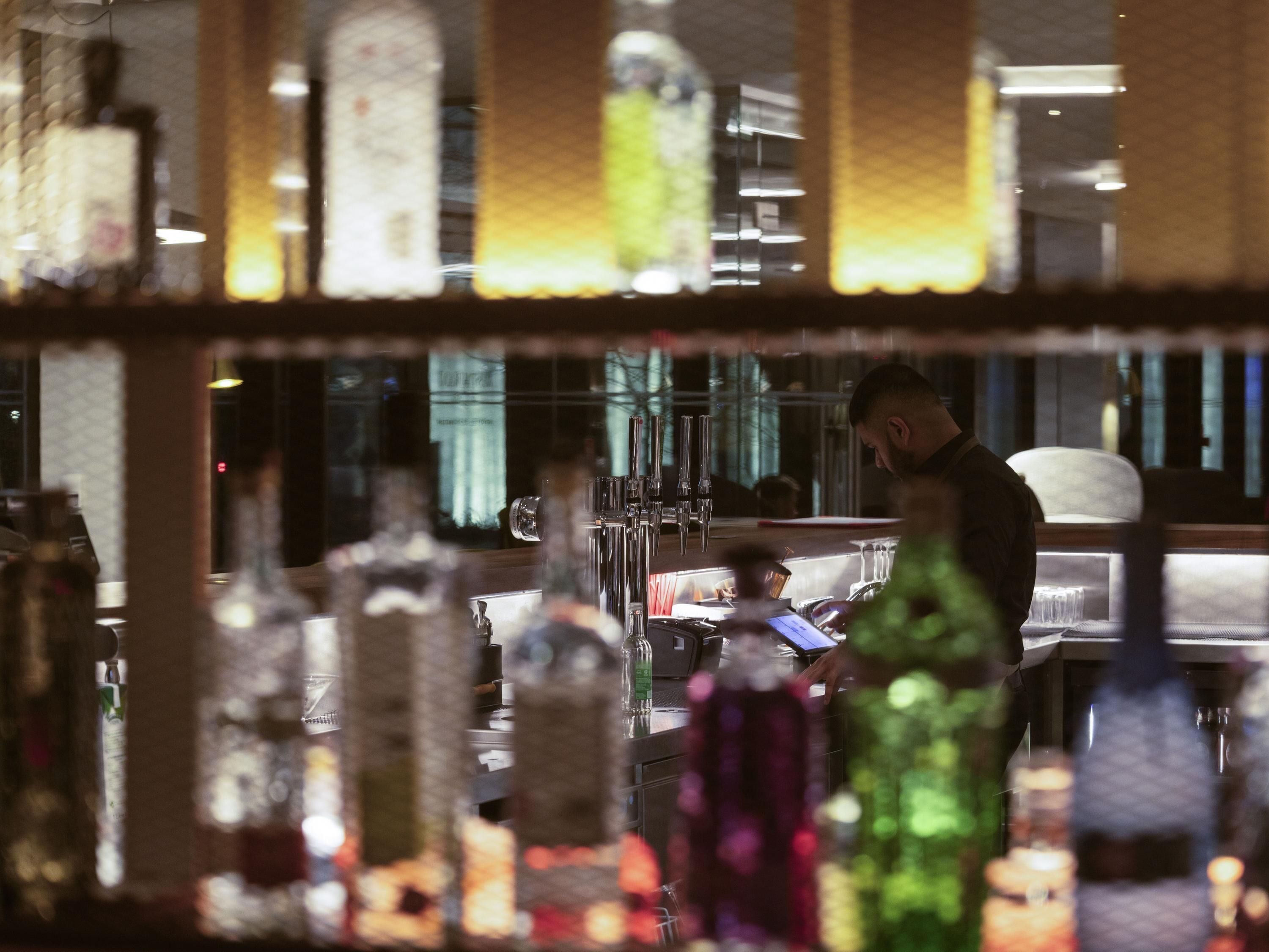 Una zona de bar con botellas y vasos, una persona trabajando detrás de la barra y el reflejo de alguien en un espejo.