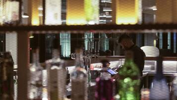 Una zona de bar con botellas y vasos, una persona trabajando detrás de la barra y el reflejo de alguien en un espejo.