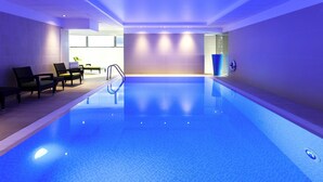 An indoor swimming pool with clear water, a diving board, and lounge chairs. - Novotel London Paddington (London)