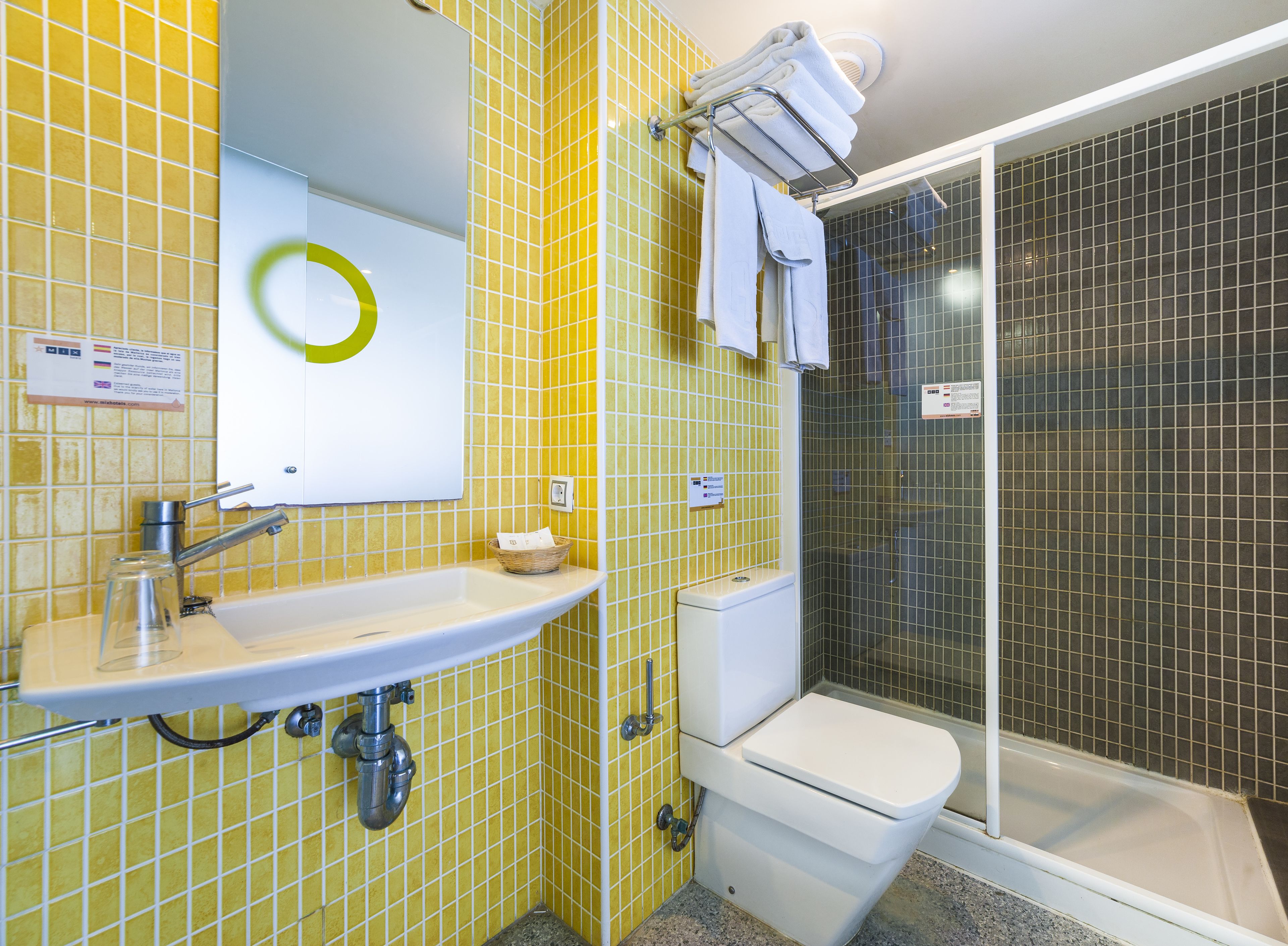 Un baño con paredes de azulejos amarillos, un lavamanos blanco, un espejo, un inodoro y una zona de ducha.