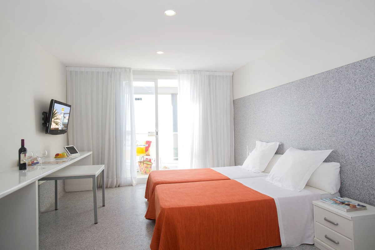 Quarto de hotel com duas camas, uma escrivaninha, televisão e janela com cortinas.