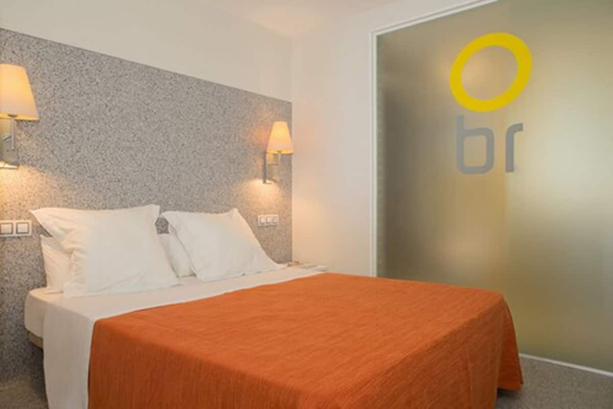 Quarto de hotel com cama, duas luminárias fixadas na parede e porta com logotipo.