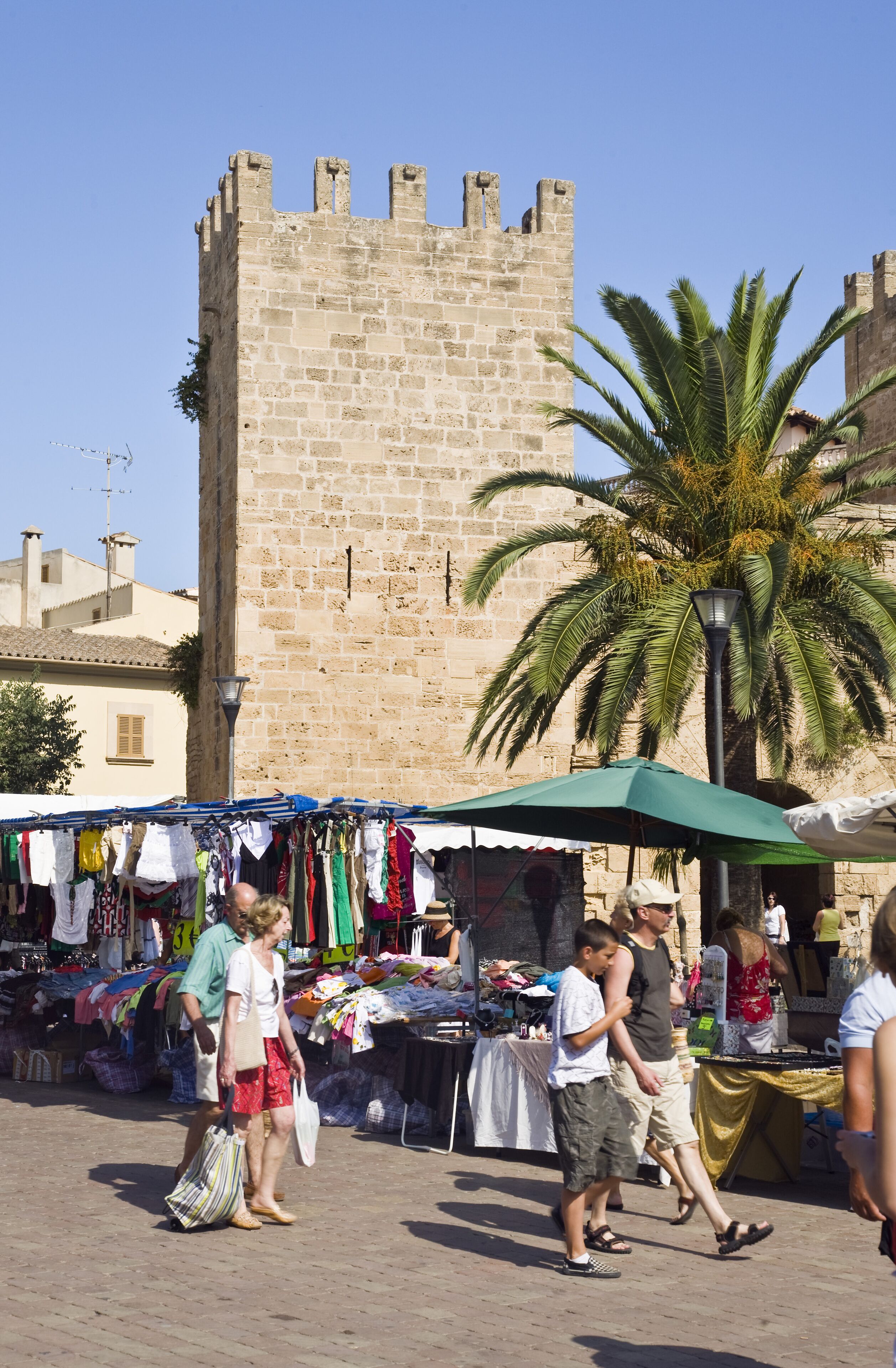 A bustling outdoor market with stalls selling clothes, set against a backdrop of a historic stone tower.