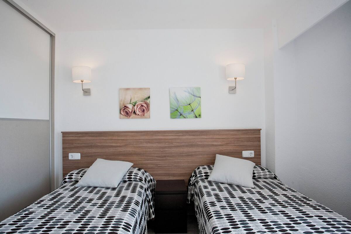 Ein Doppelzimmer mit zwei Betten, jeweils mit einem gemusterten Überwurf, einem Holzkopfstück und je einer Wandlampe.