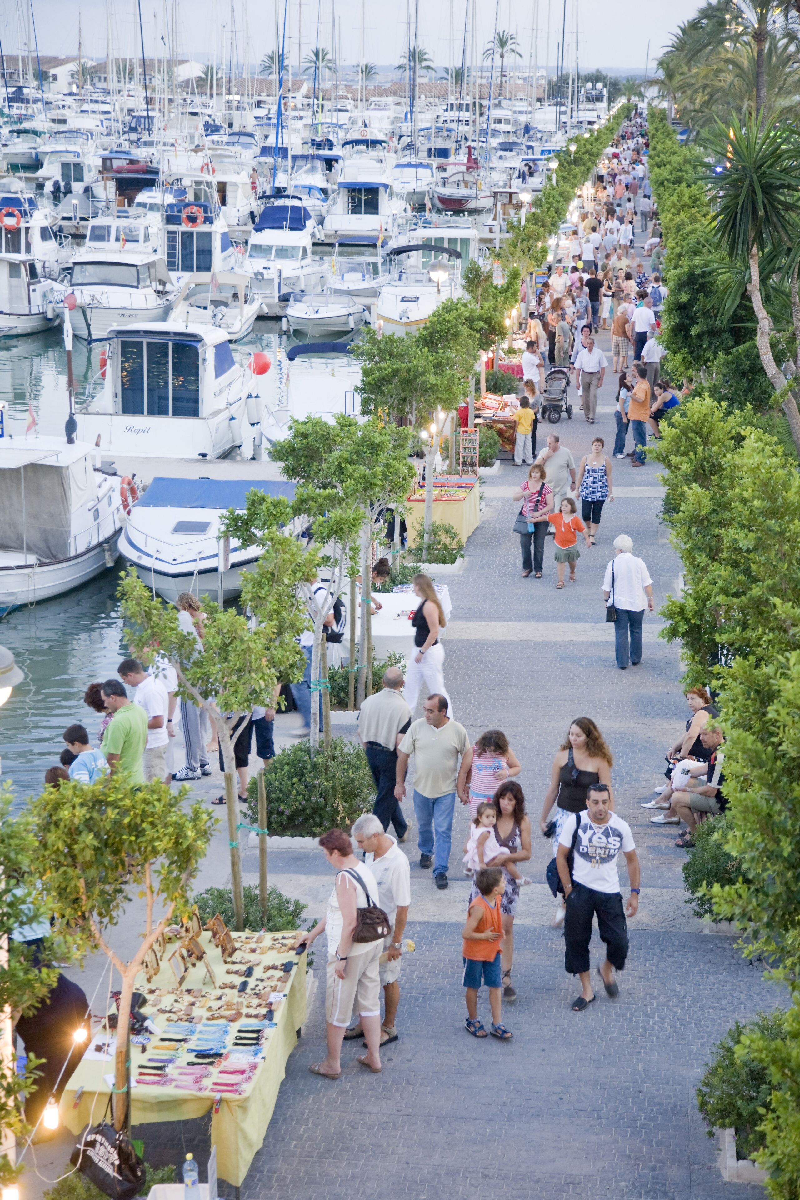 A marina with boats docked and a pedestrian walkway lined with trees and vendors.