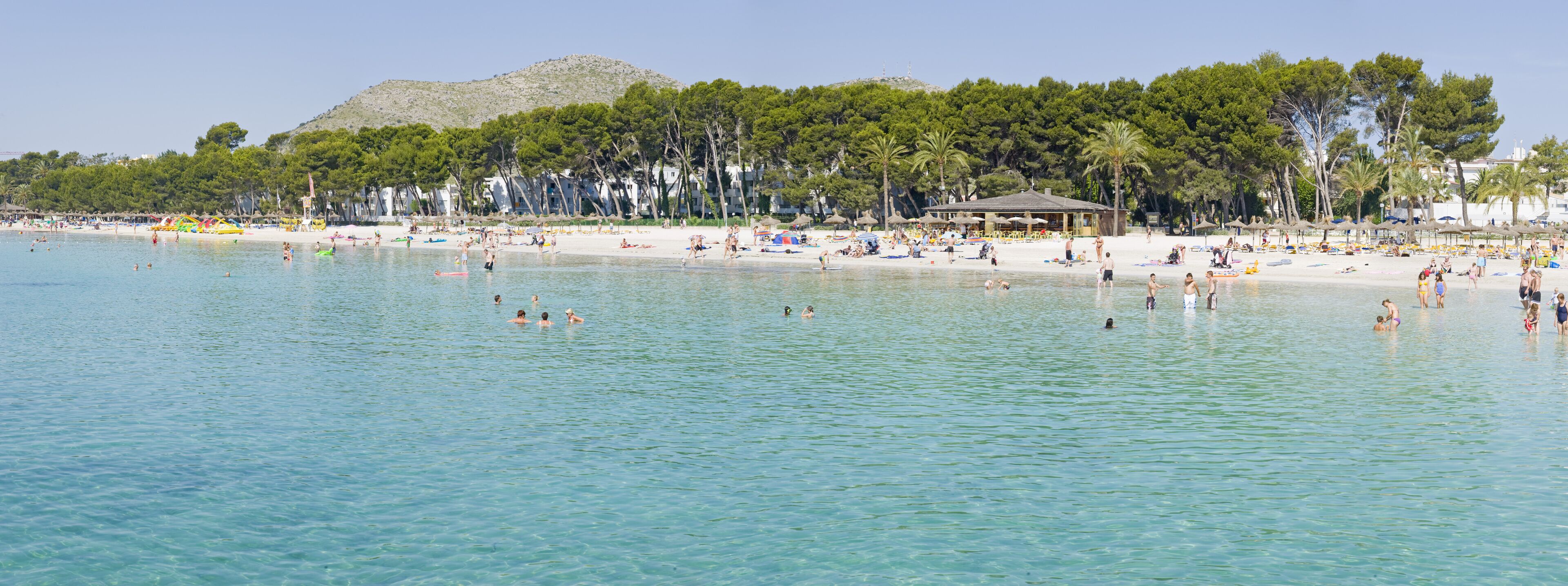 Una playa con agua turquesa cristalina, gente nadando y tomando el sol, y un telón de fondo de árboles y colinas.