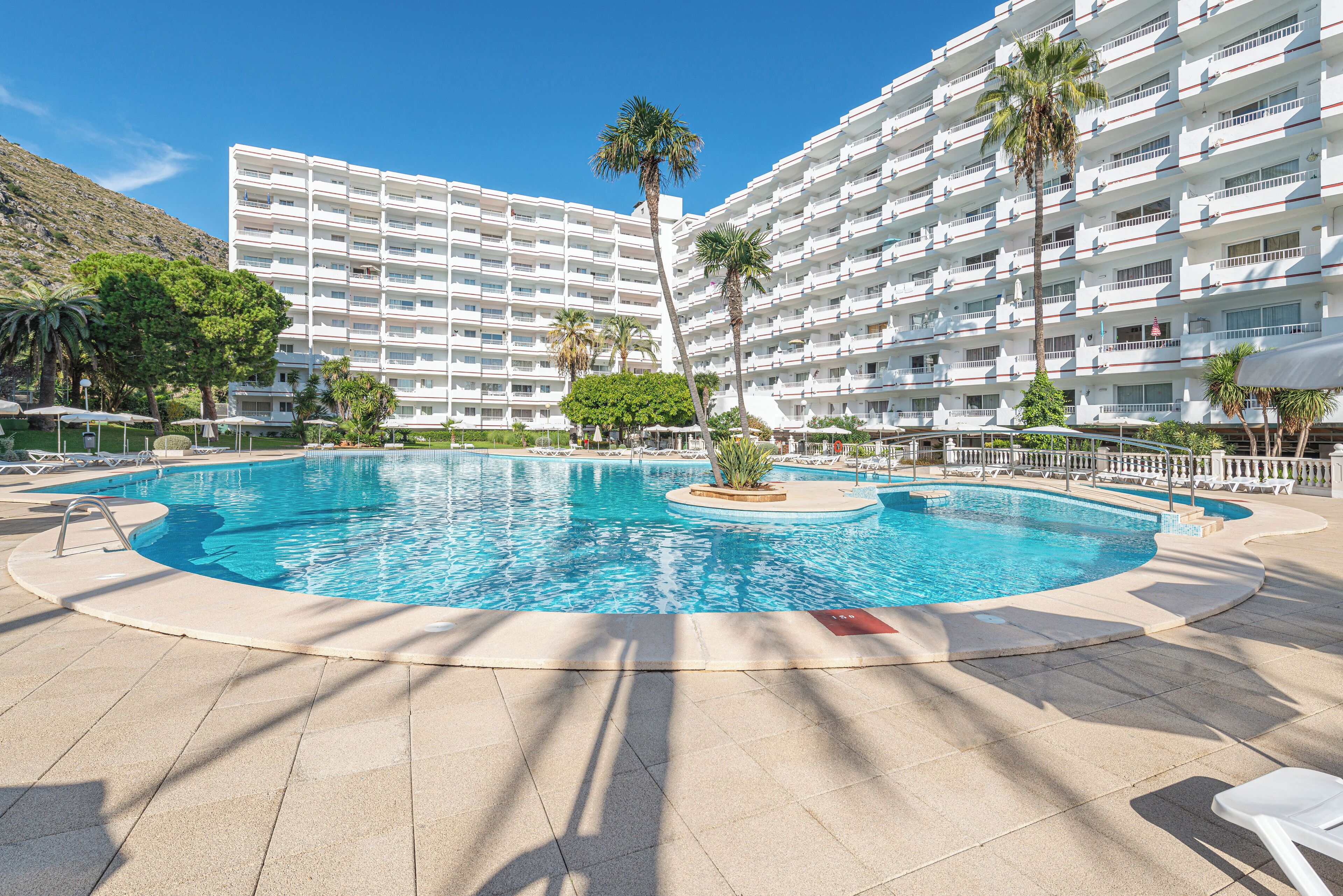 A large swimming pool surrounded by a white building with multiple balconies, palm trees, and sun loungers.