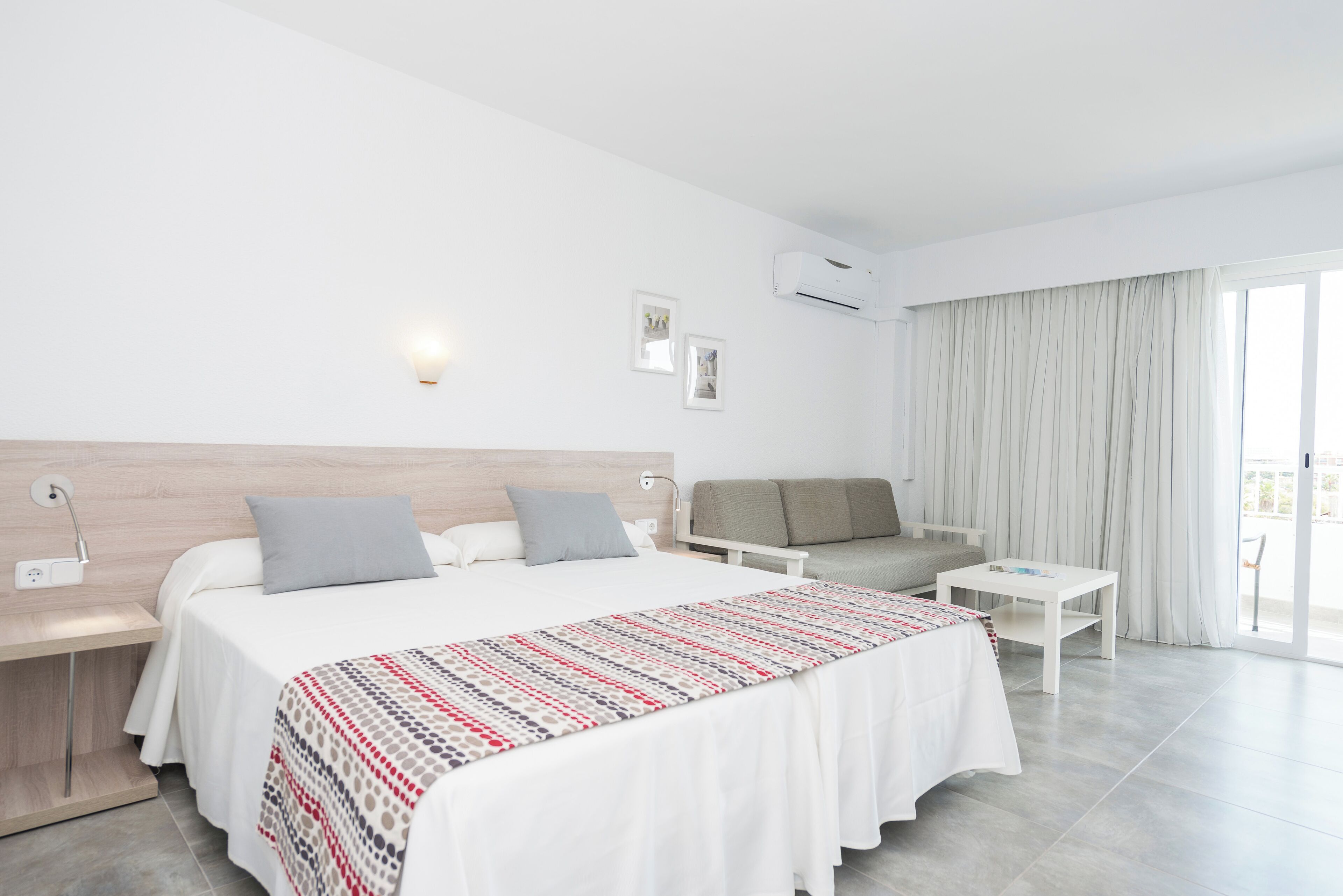 Een moderne hotelkamer met een groot bed, een slaapbank, een tafeltje en een balkon met gordijnen.