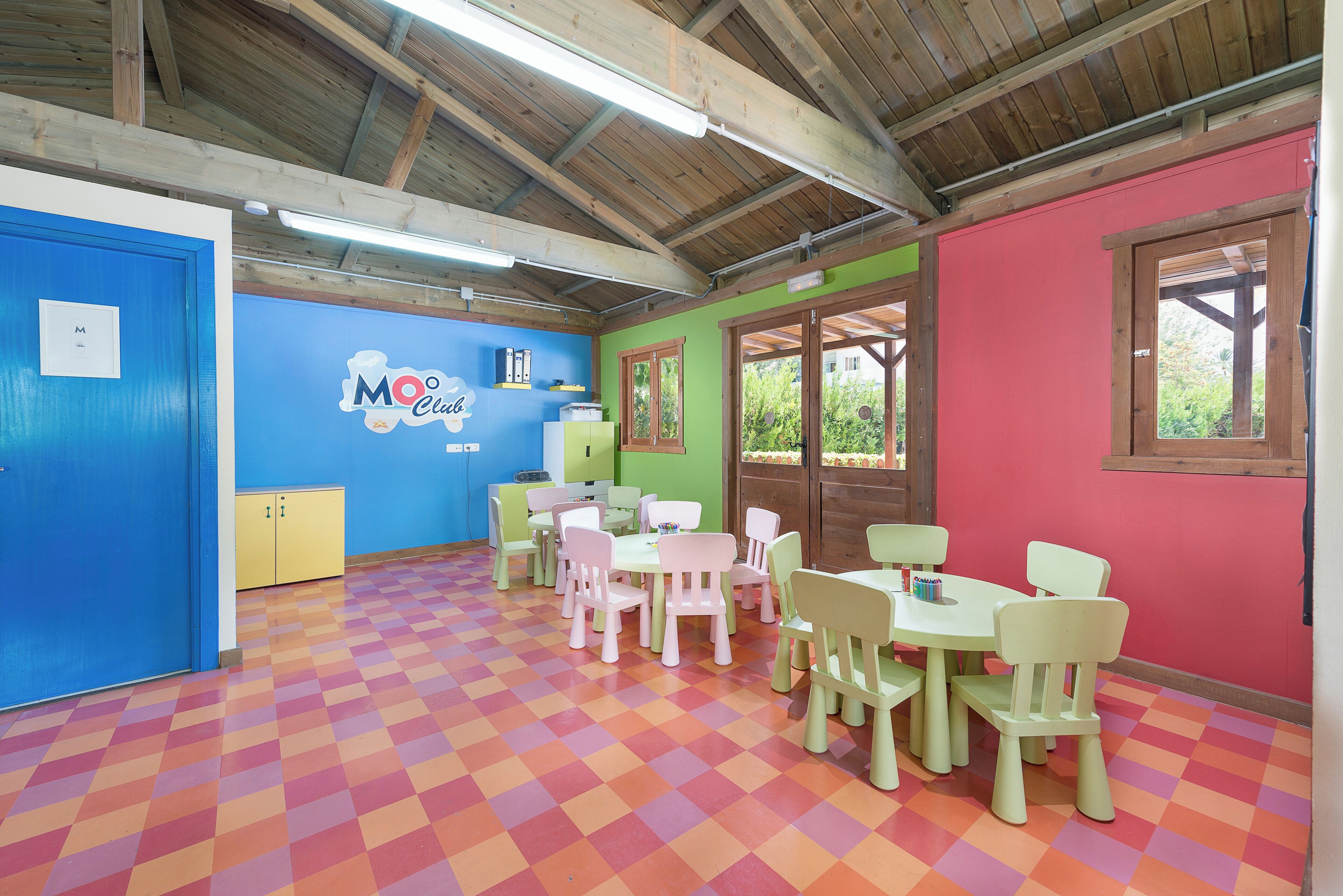 Une salle de jeux pour enfants avec un sol à carreaux multicolores, une porte bleue, un meuble jaune et une table avec des chaises.