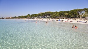 A beach with clear shallow water, people swimming and sunbathing, and a backdrop of trees and buildings. - Apartamentos Siesta I (Alcúdia)