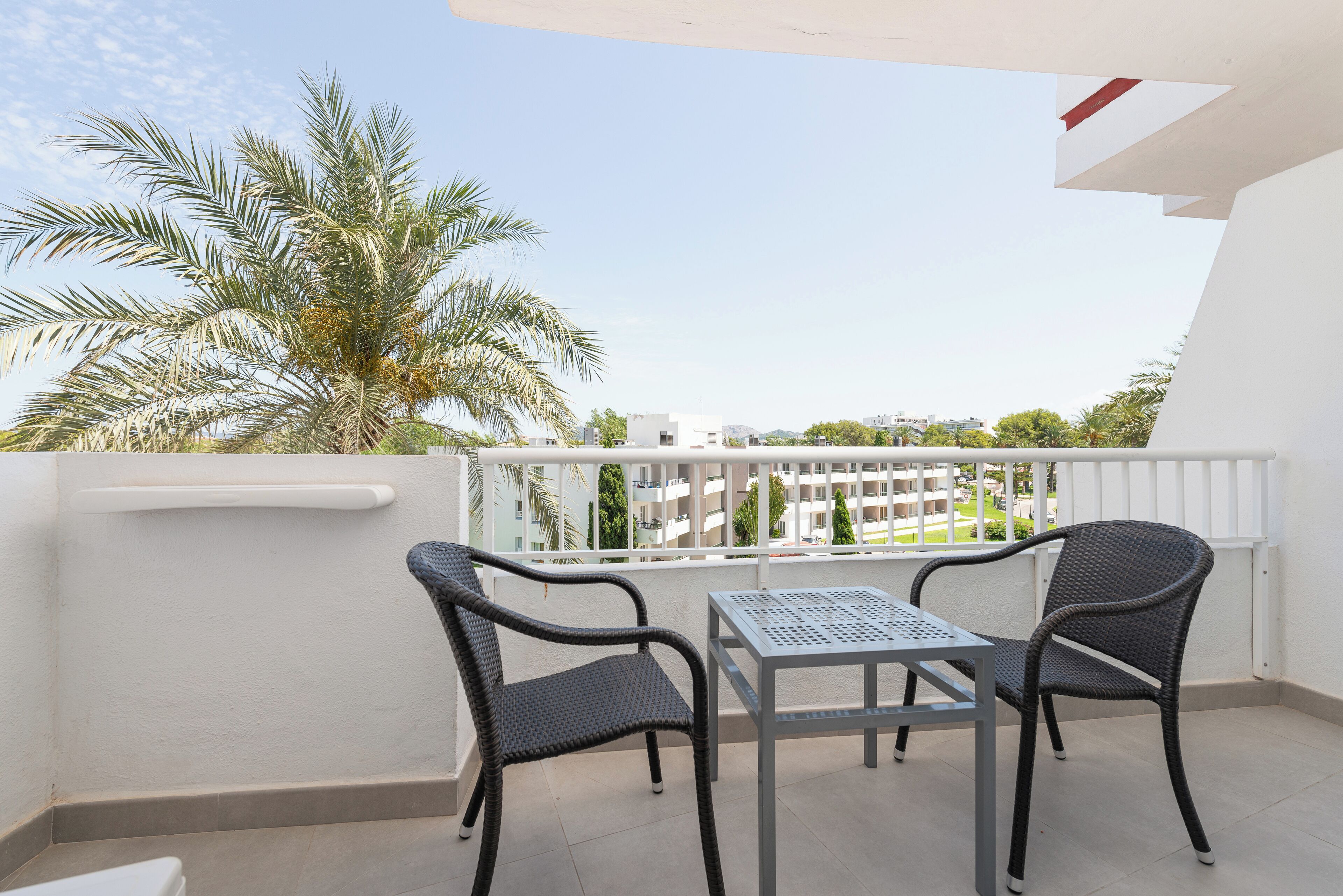 Un balcón con dos sillones de mimbre y una mesa de metal, con vistas a una zona residencial con palmeras y edificios.