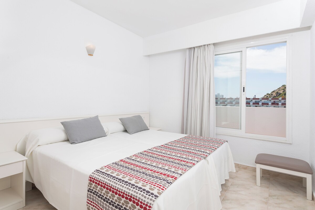 Una habitación de hotel con cama, una banca, una mesita de noche y una ventana con vistas a edificios y colinas.