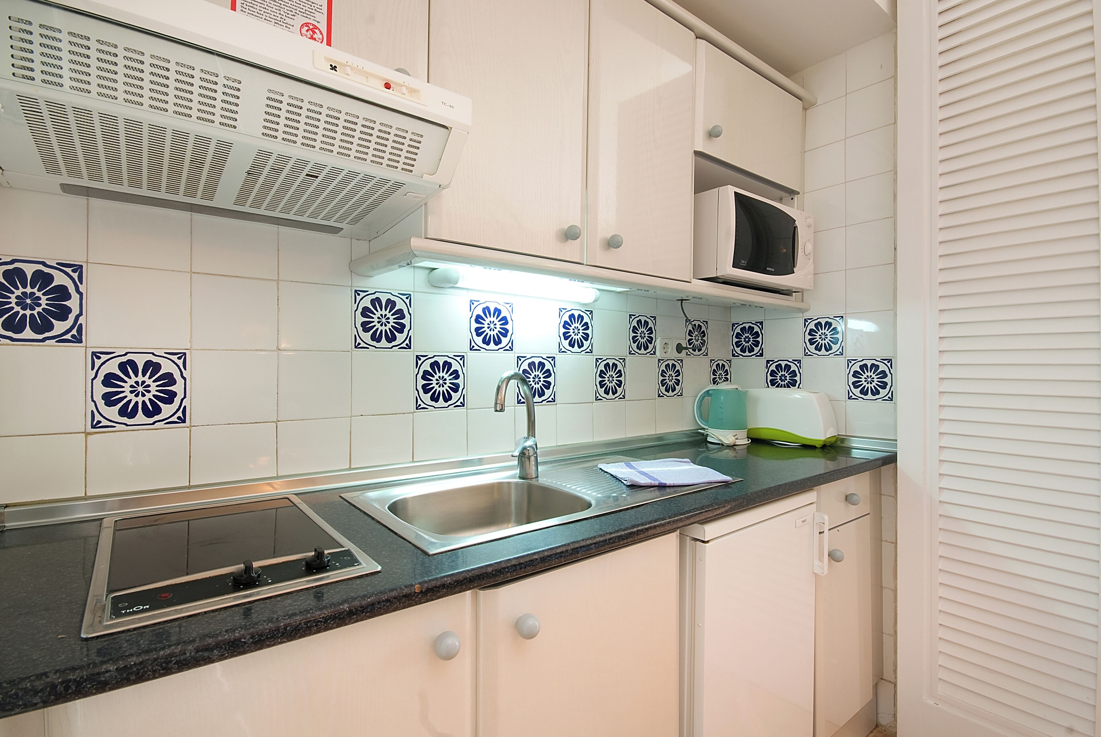 A kitchen with white cabinets, a black countertop, a stainless steel sink, a microwave, and decorative blue and white tiles.