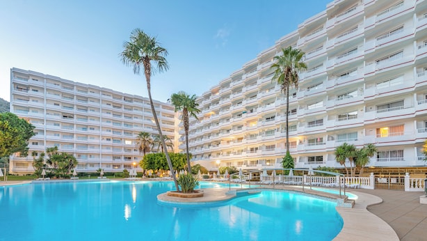 A large swimming pool surrounded by a multi-story hotel with balconies and palm trees. - Apartamentos Siesta I (Alcúdia)