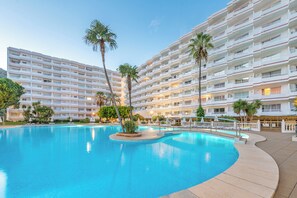 A large swimming pool surrounded by a multi-story hotel with balconies and palm trees. - Apartamentos Siesta I (Alcúdia)