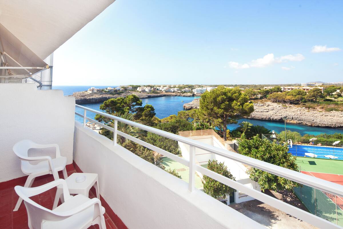 A balcony with white chairs, a tennis court, and a coastal view.