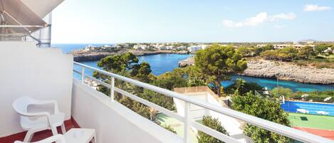 A balcony with white chairs, a tennis court, and a coastal view.