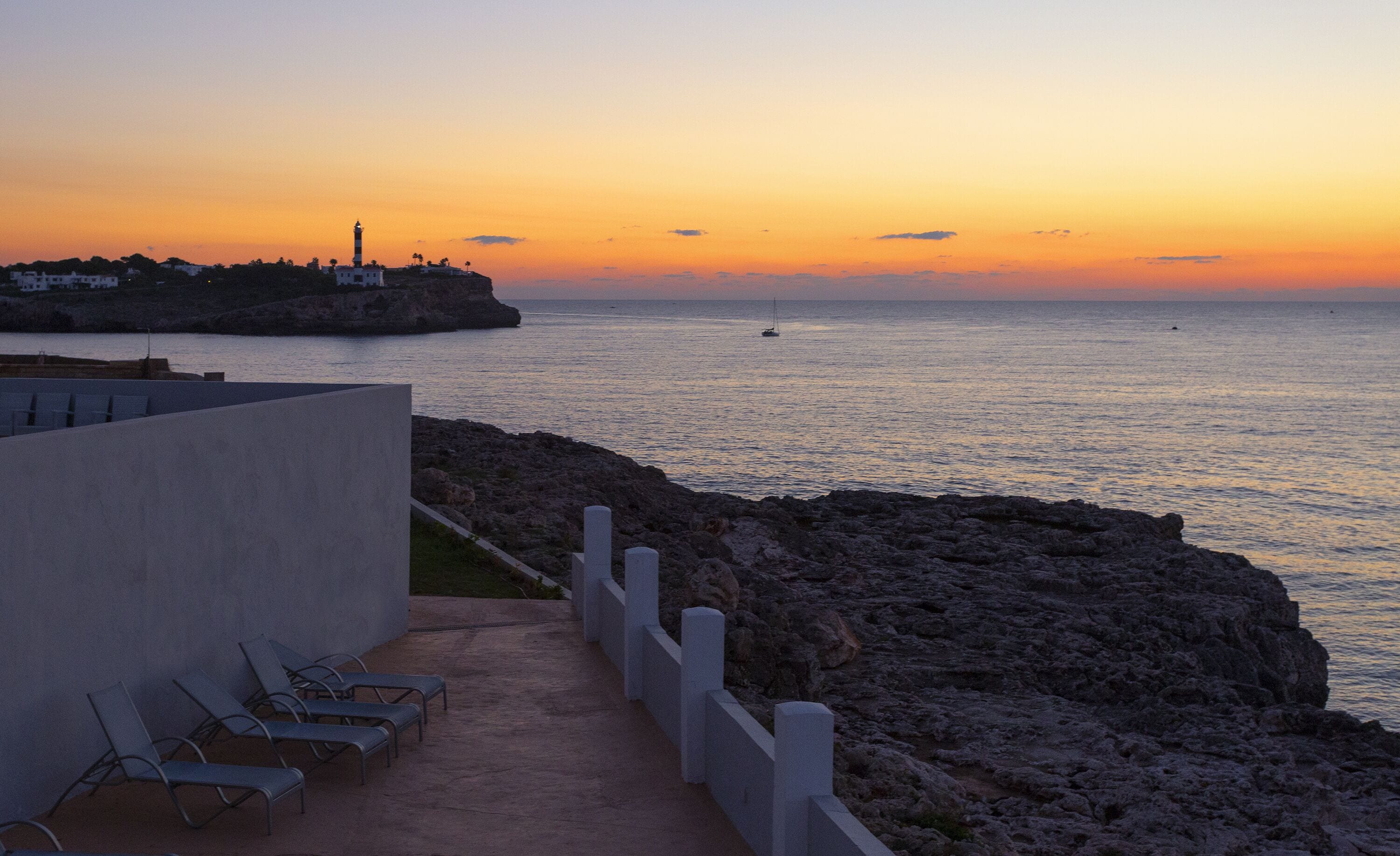Vista sulla costa con un litorale roccioso, un faro e una fila di sedie a sdraio vuote.
