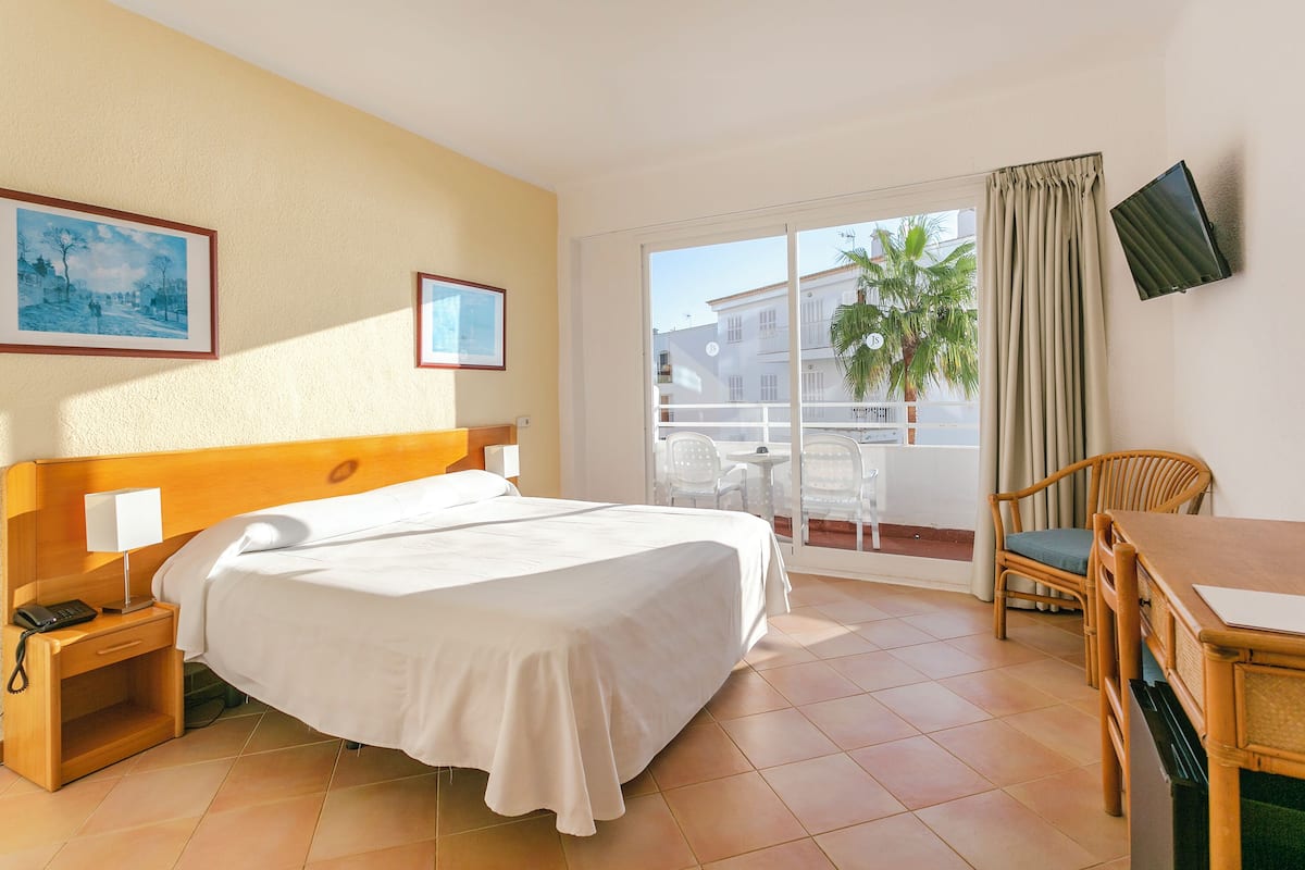 A hotel room with a bed, a desk with a chair, a TV, and a balcony with a table and chairs.