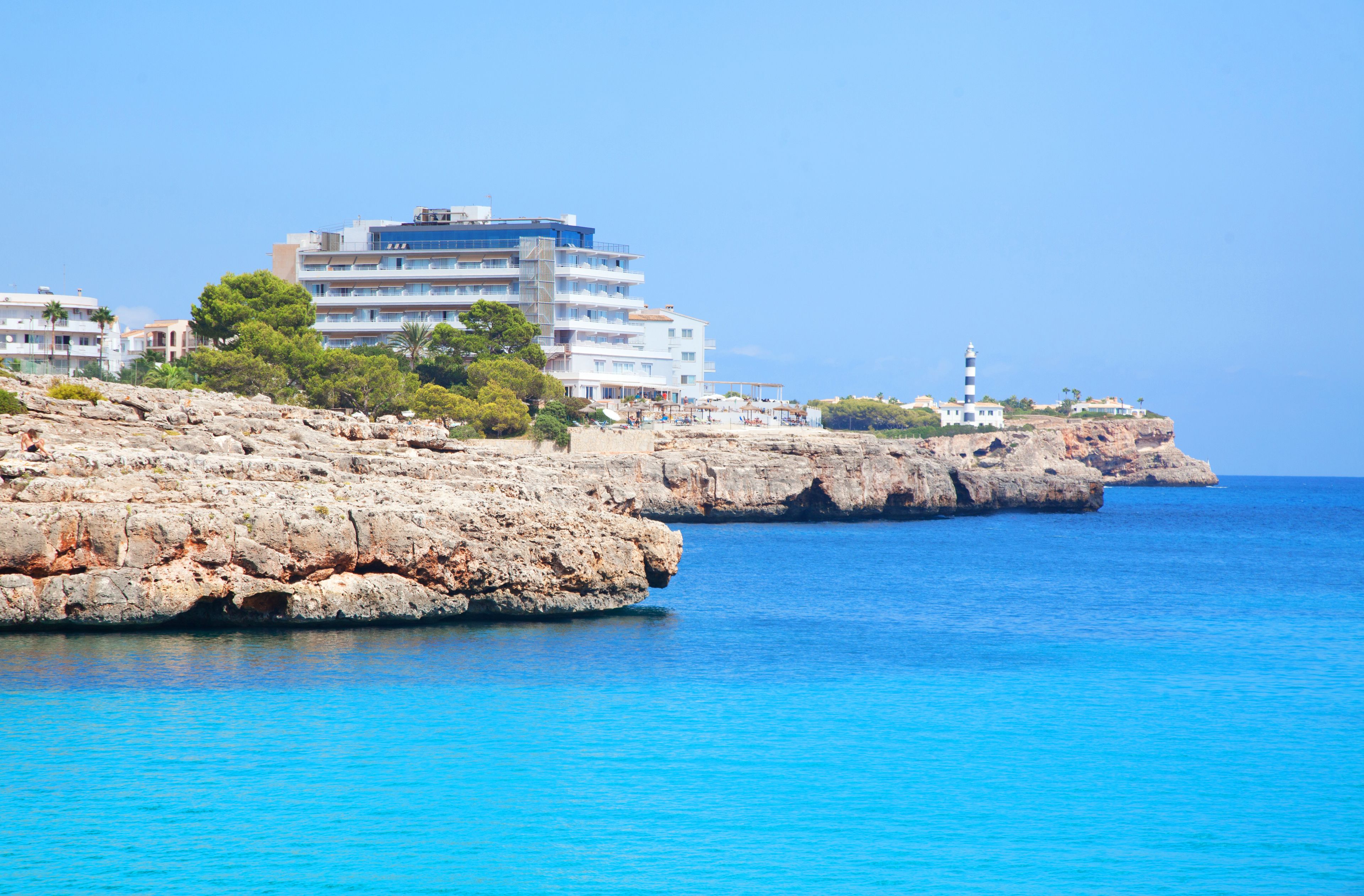 A coastal hotel with multiple floors, a rocky shoreline, and a lighthouse in the background.
