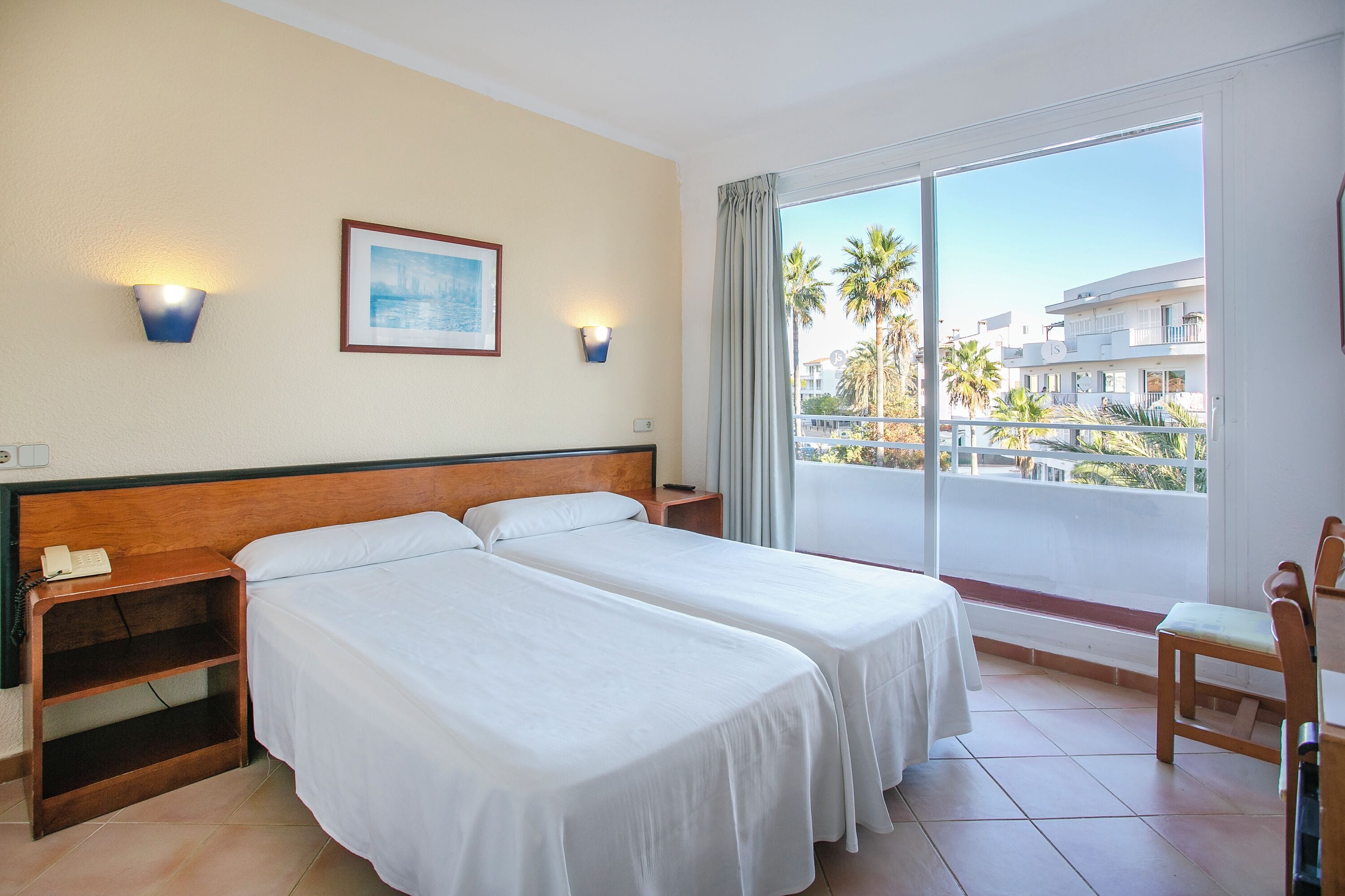 Habitación de hotel con dos camas, una mesita de noche, una silla, un ventanal con vistas a palmeras y edificios, y luces fijadas a la pared.