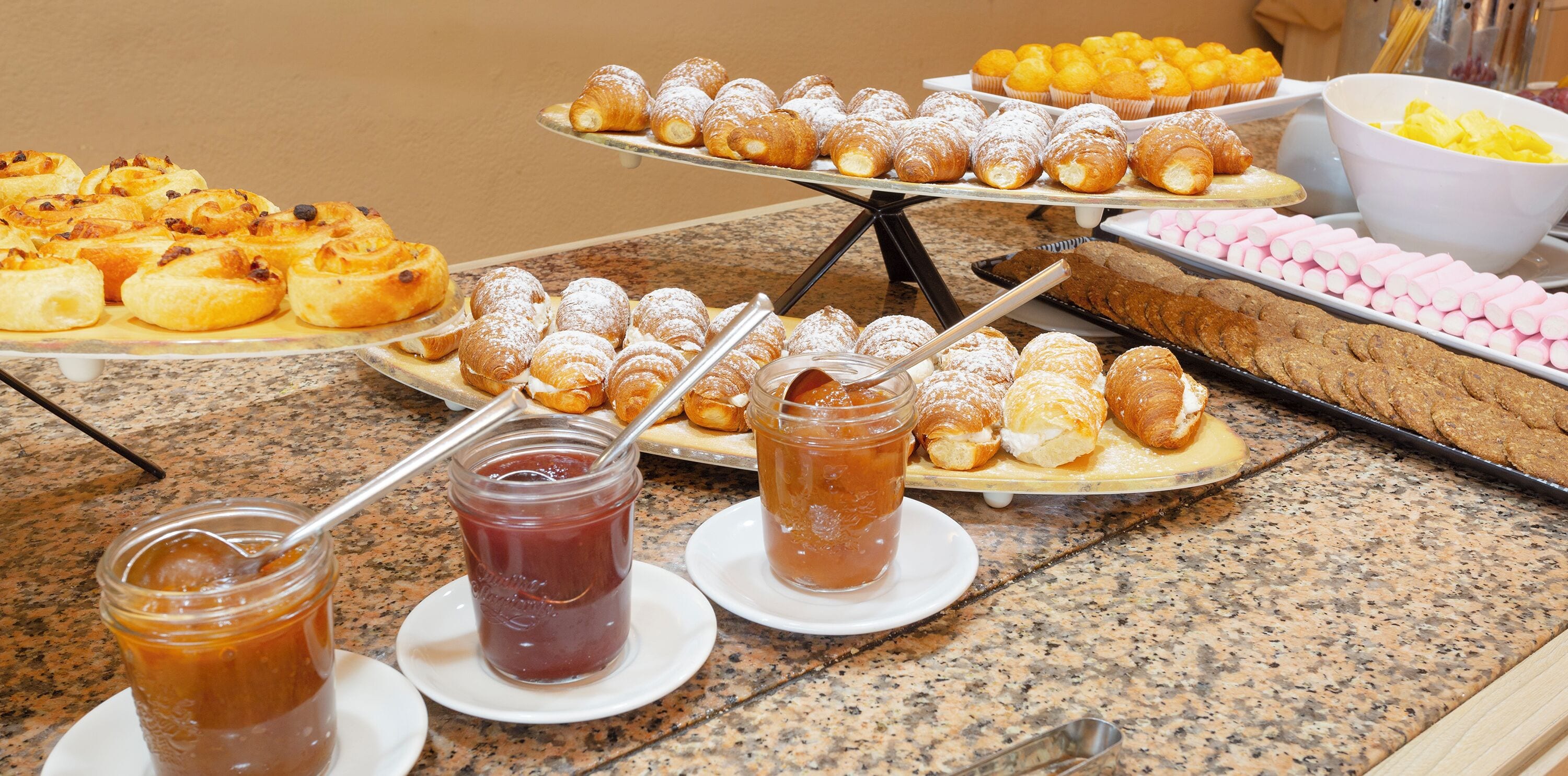 Un bufé con diversos pasteles y postres, incluyendo croissants y muffins, y dos frascos de mermelada.