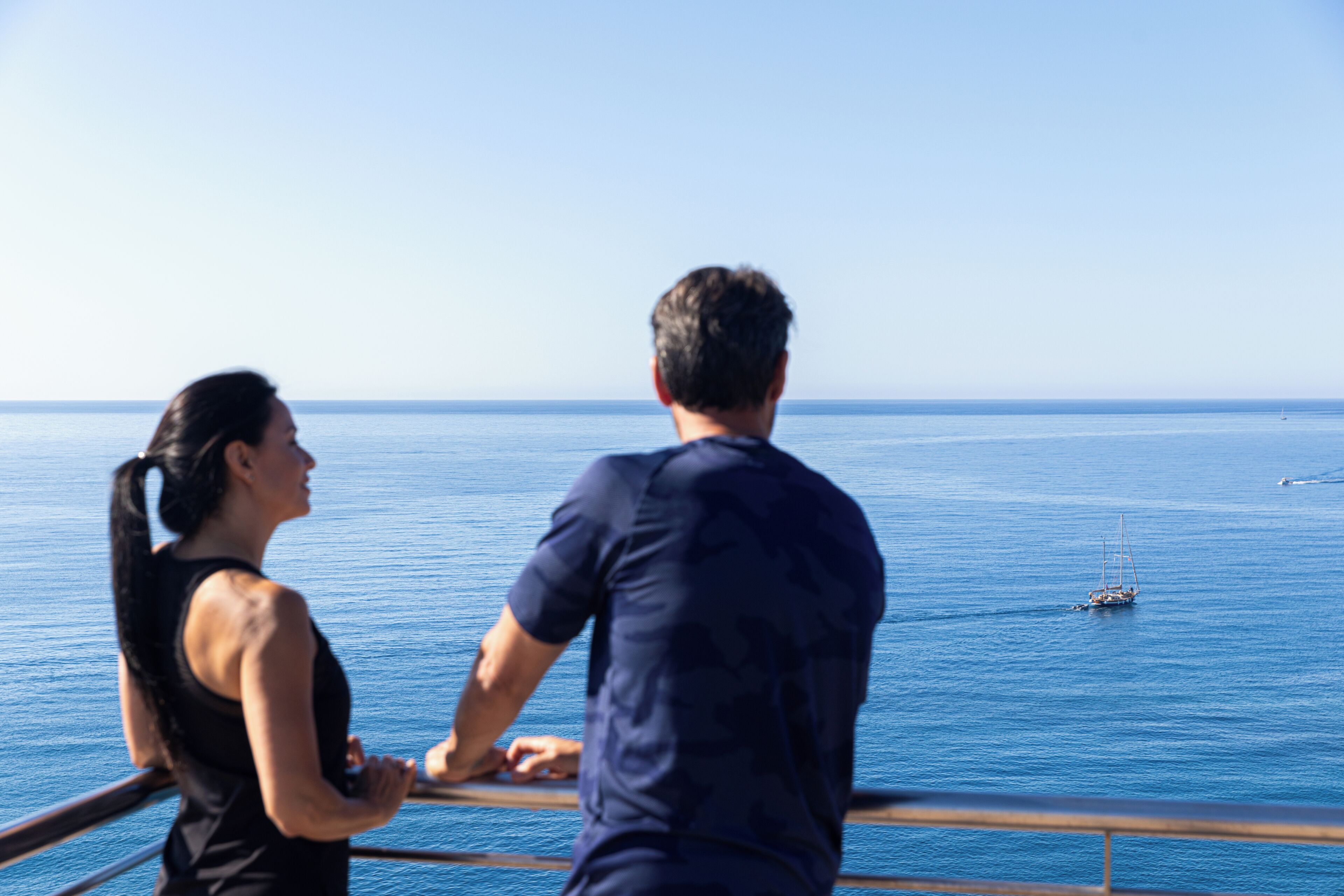 Un couple se tient sur un balcon, d’où l’on aperçoit une mer calme et, au loin, un voilier.