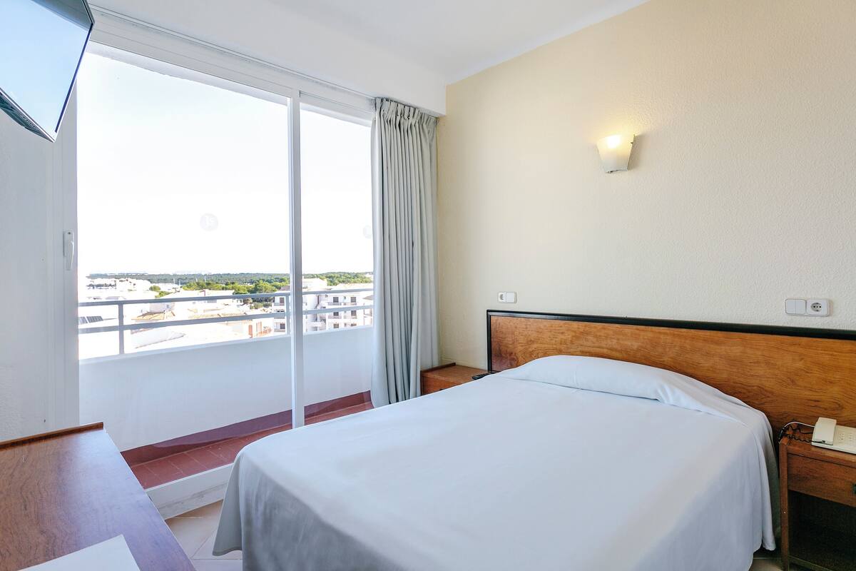 A hotel room with a bed, a bedside table with a phone, a wall-mounted light, and a view through a sliding door to an outdoor area.