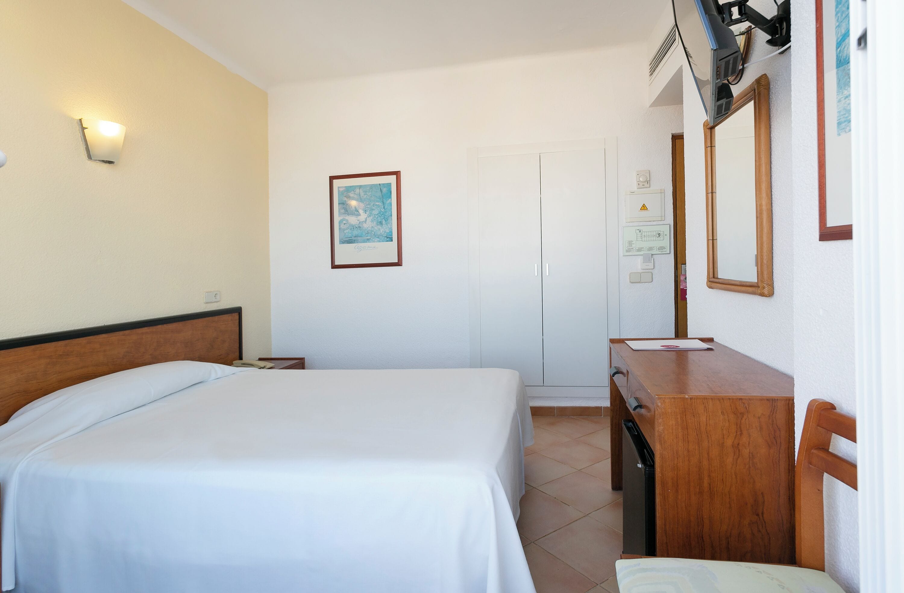 A hotel room with a bed, a desk, a chair, a mirror, and a wall-mounted light.