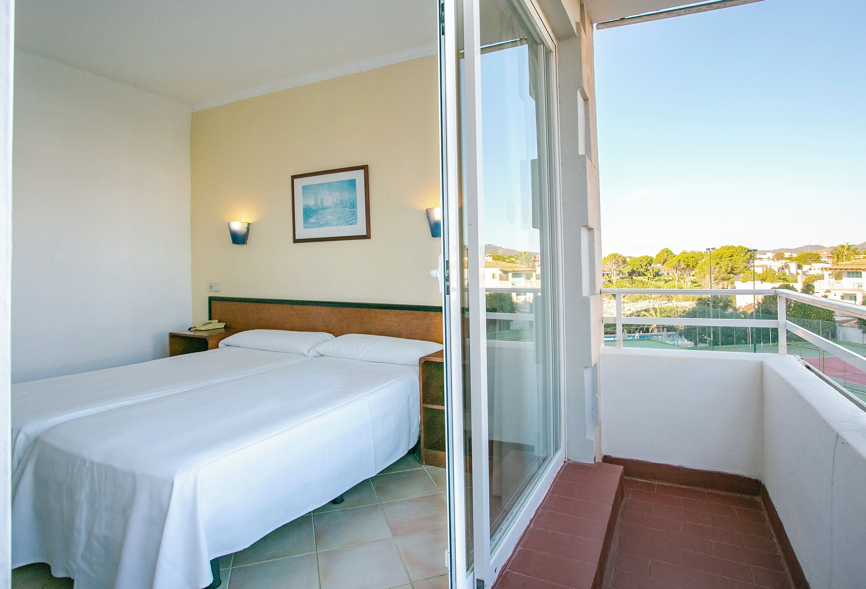 A hotel room with a bed, a nightstand, a framed picture on the wall, and a balcony with a view of the city.