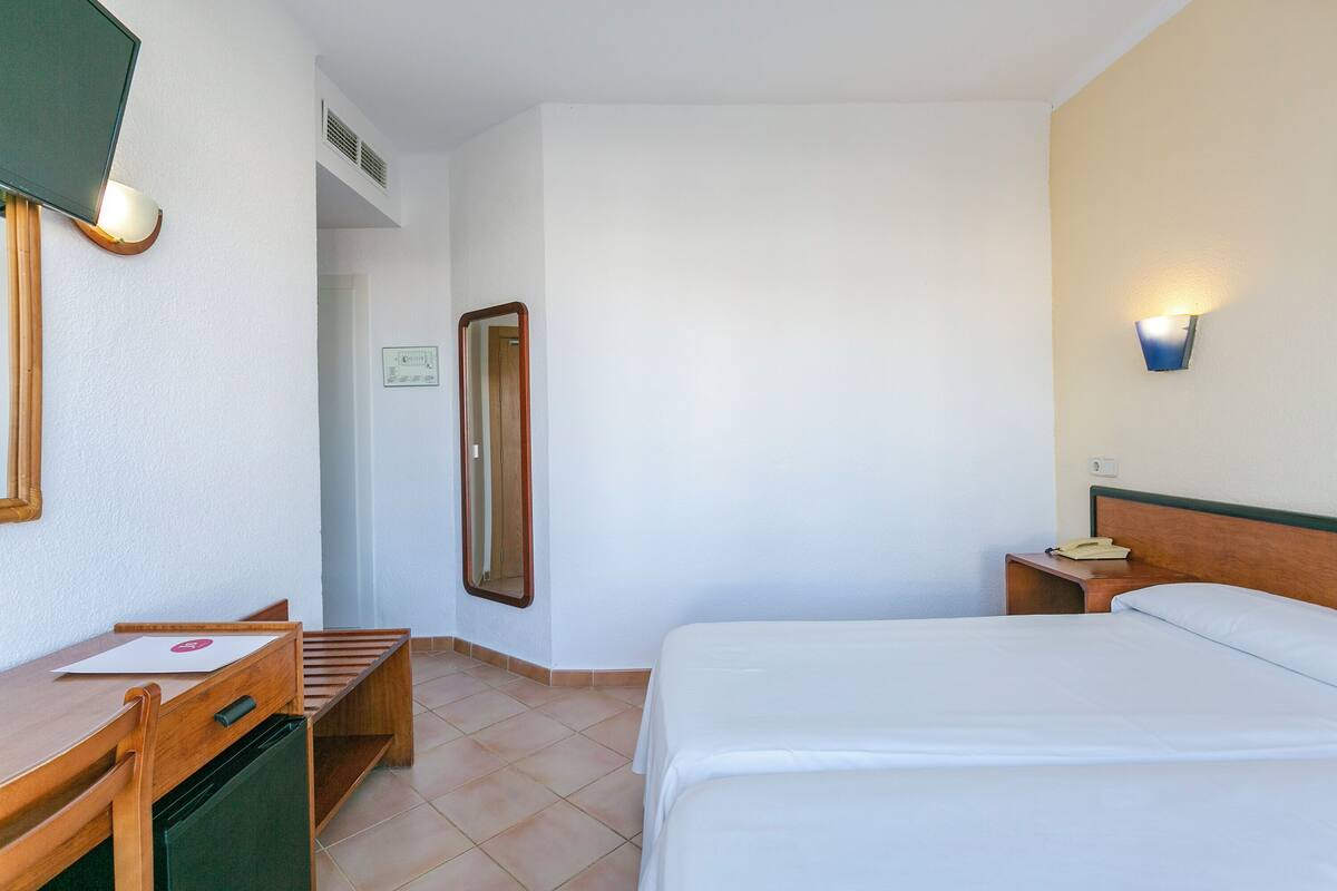 A hotel room with a bed, a desk with a chair, a mirror, a nightstand with a phone, and a wall-mounted light.
