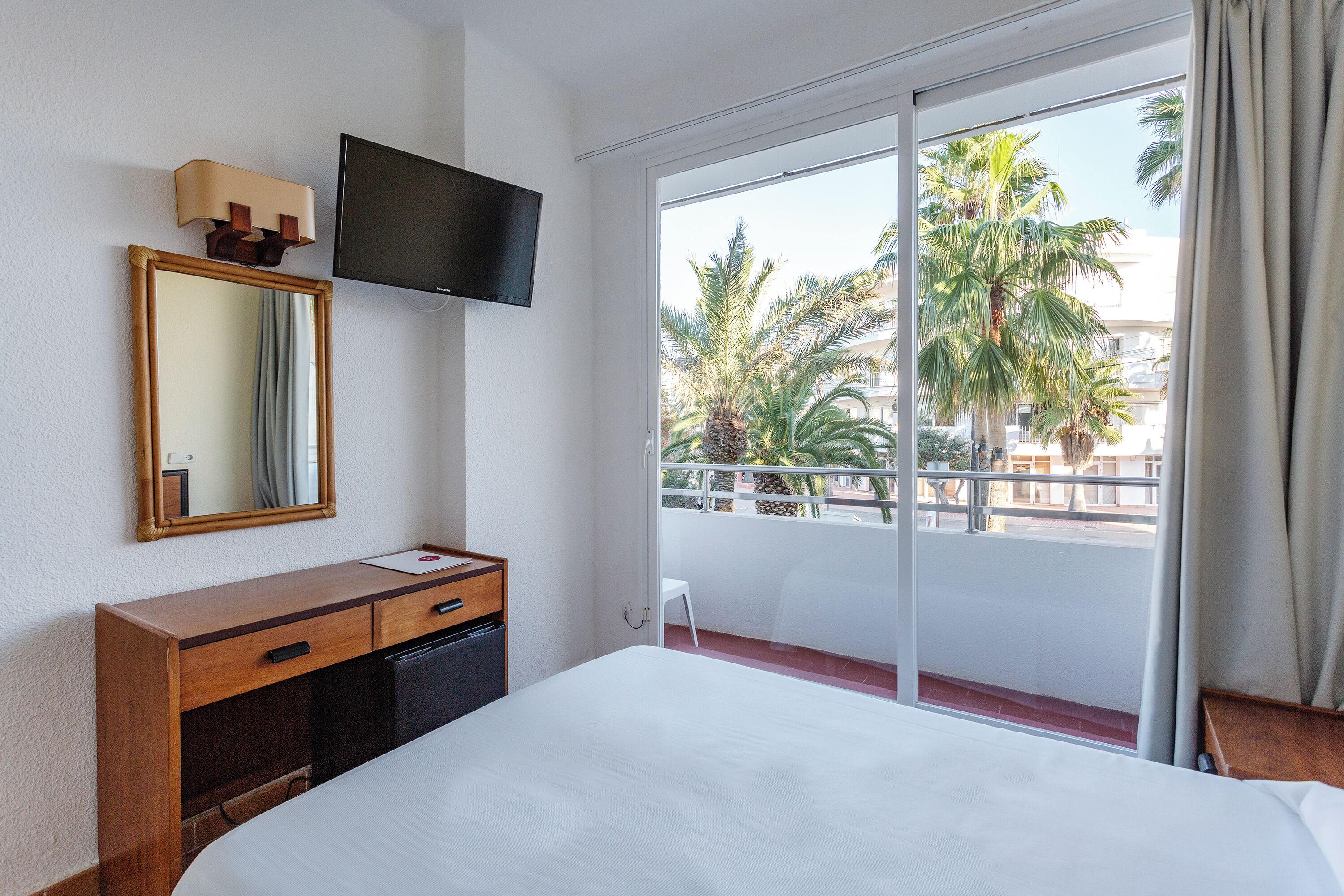 Habitación de hotel con cama, televisión, escritorio, espejo y balcón con vista a palmeras.