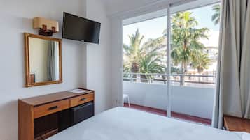 Habitación de hotel con cama, televisión, escritorio, espejo y balcón con vista a palmeras.