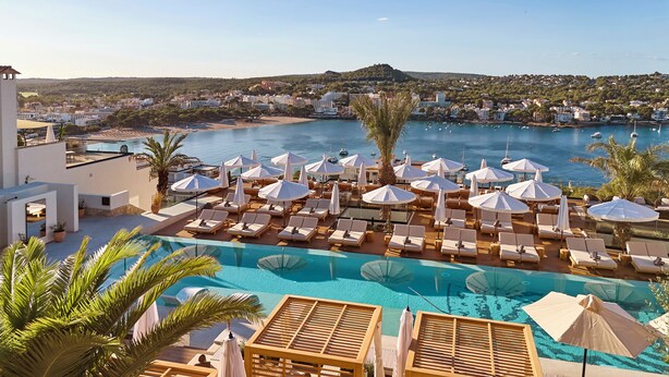 Un espace piscine aménagé dans un complexe hôtelier, avec des chaises longues et des parasols, donnant sur une étendue d’eau où se trouvent des bateaux.