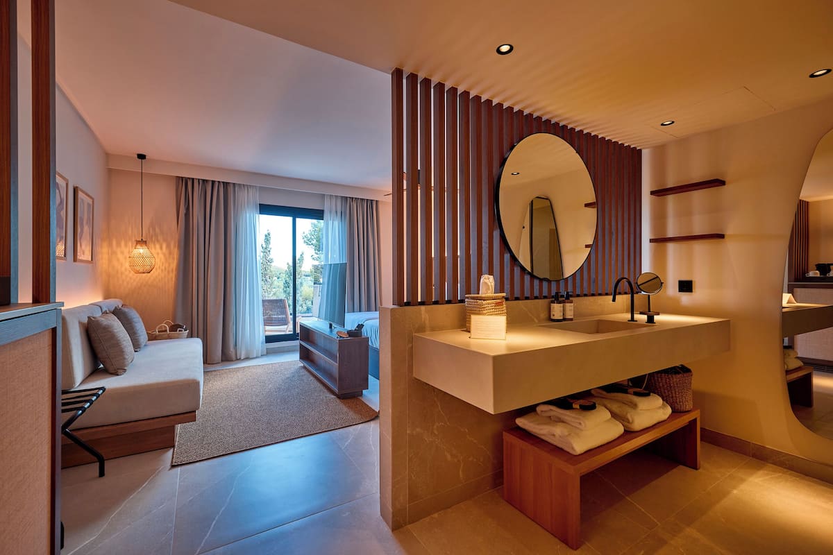A modern hotel room with a sofa, a large round mirror, a built-in sink, and a view of the outdoors.
