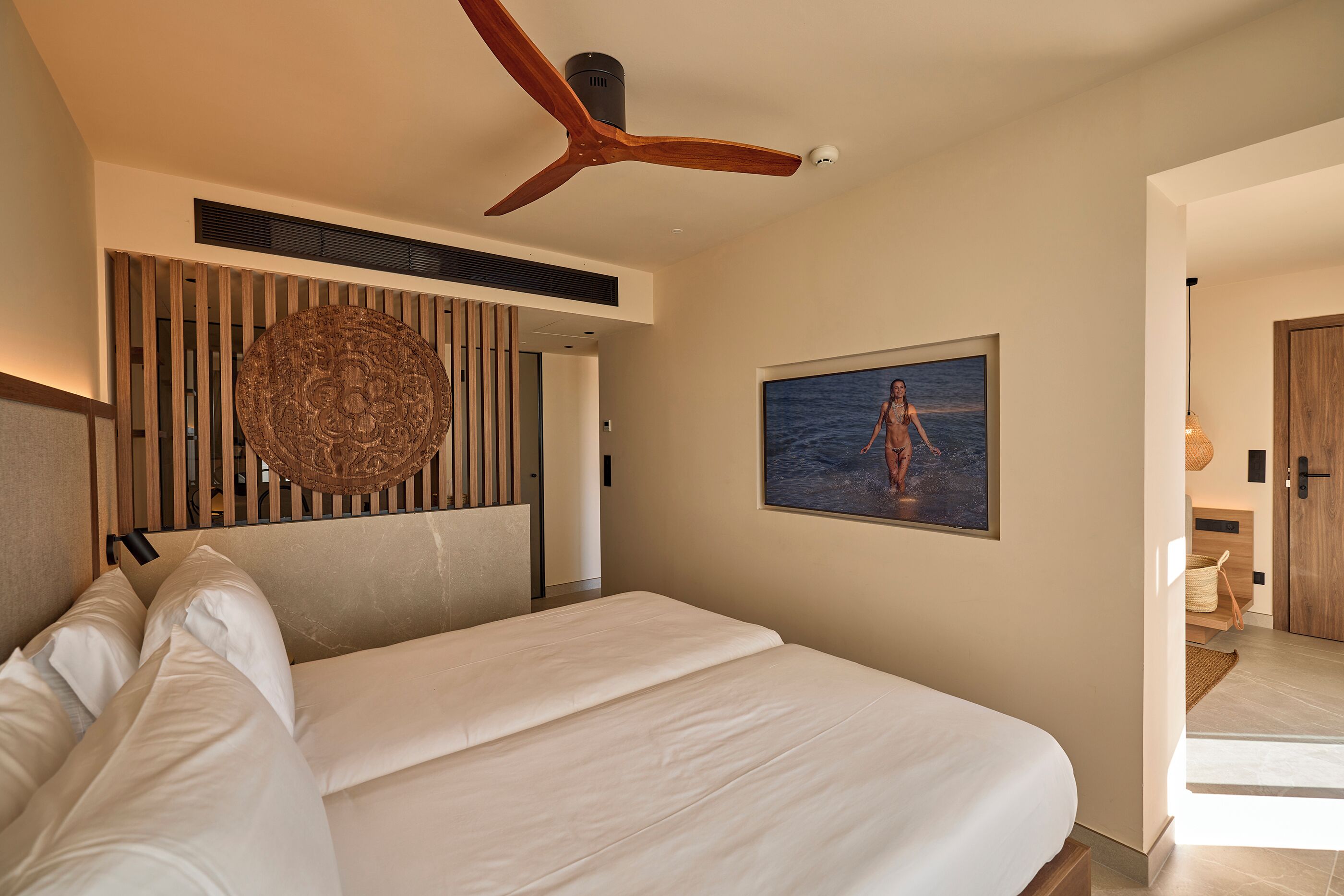 A modern hotel room with a large bed, a ceiling fan, a wall-mounted artwork, and a view of the sea through the window.