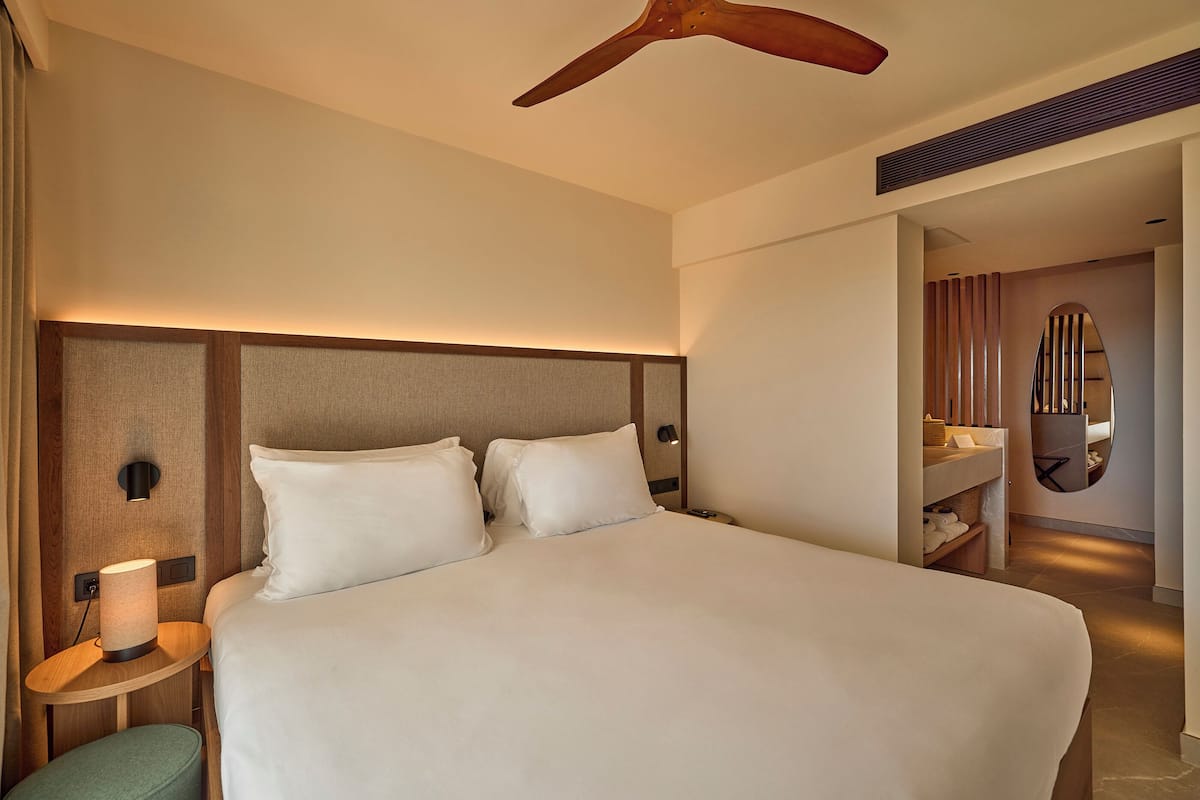 A modern hotel room with a large bed, a bedside table with a lamp, a ceiling fan, and a bathroom visible through an open door.