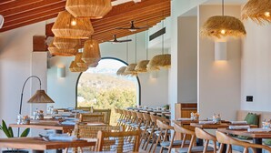 Un restaurant avec des tables et des chaises en bois, des luminaires suspendus en fibres tressées et une vue sur les arbres à travers de grandes fenêtres.