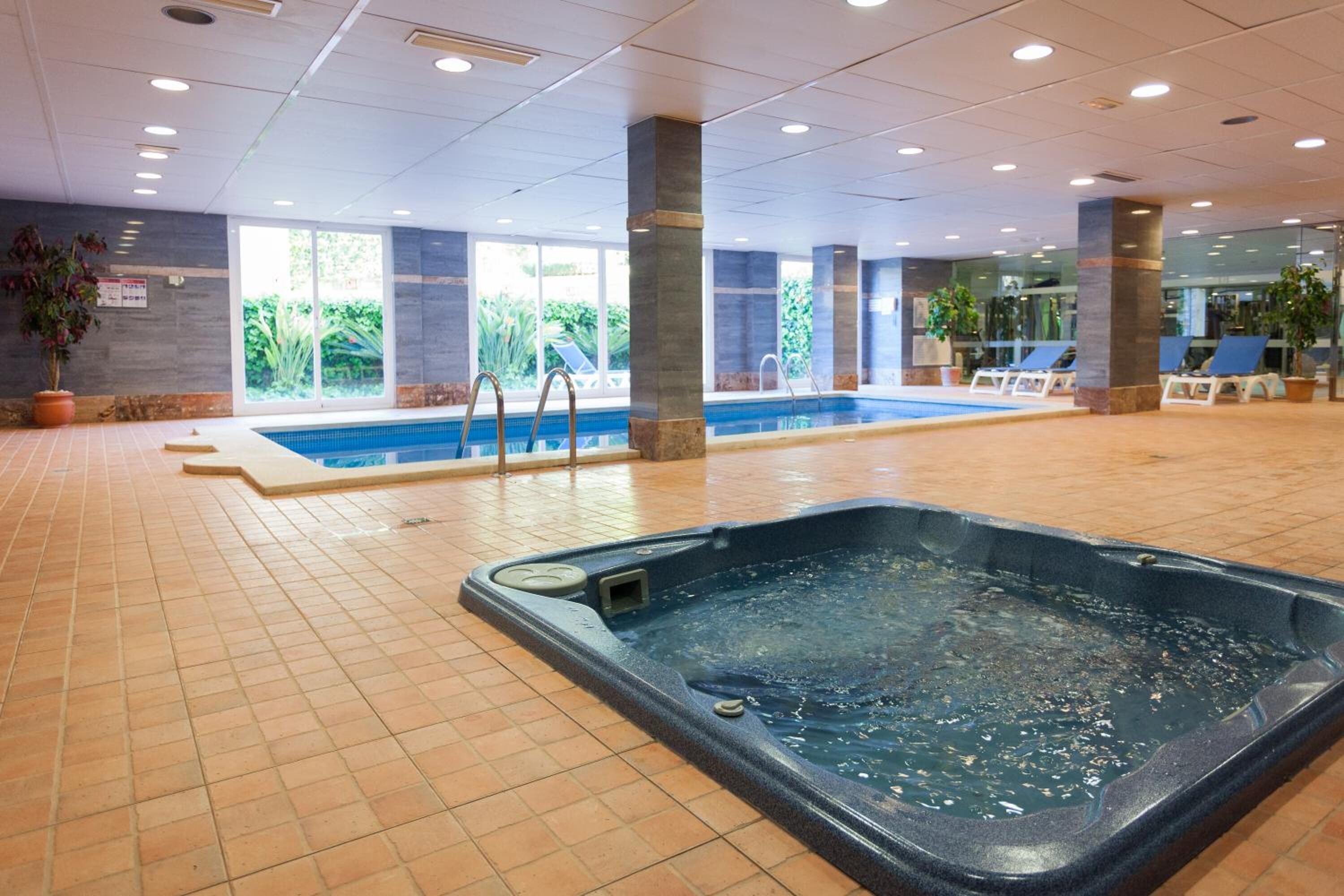 Un área de piscina interior con jacuzzi, suelo de baldosas y sillones de descanso.