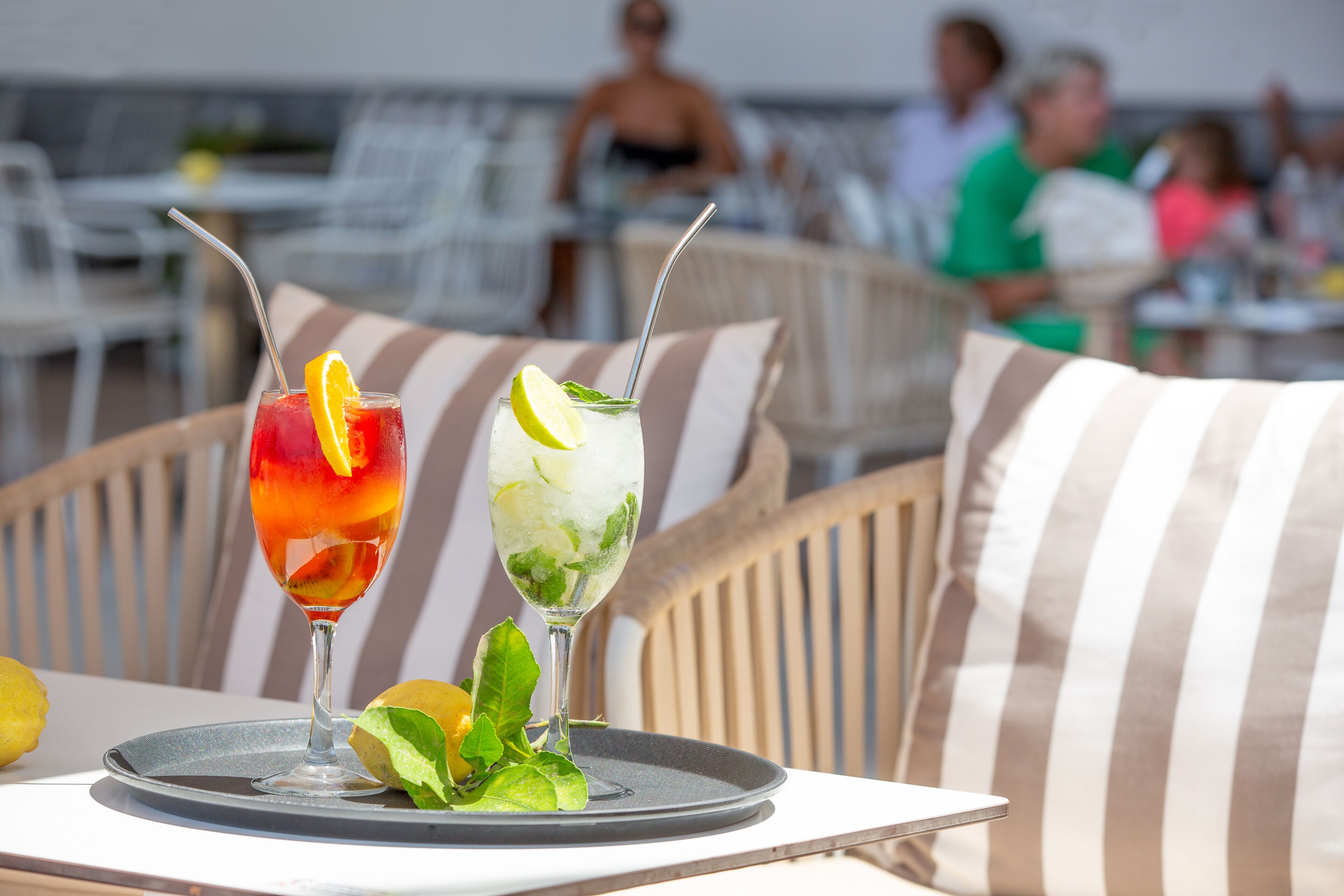 Two cocktails on a tray with a lemon slice garnish, placed on a table with striped cushions.