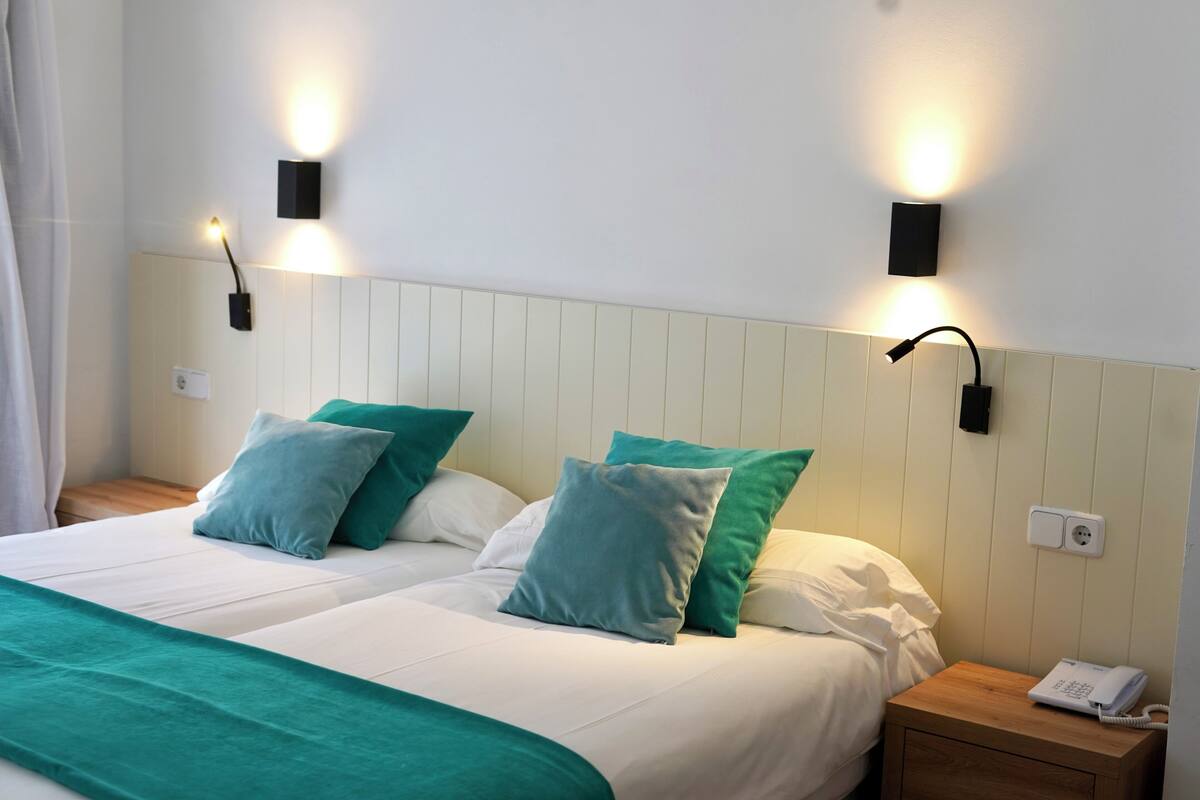 Een hotelkamer met twee bedden, blauwgroene beddengoed, een houten nachtkastje met een telefoon en wandlampen.
