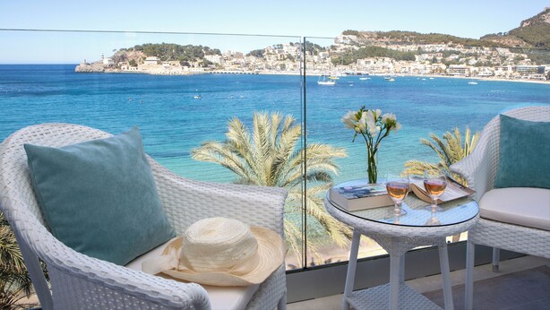 Un balcon avec des fauteuils en osier, une petite table avec des boissons et un chapeau, d’où l’on peut admirer une ville côtière et la mer.