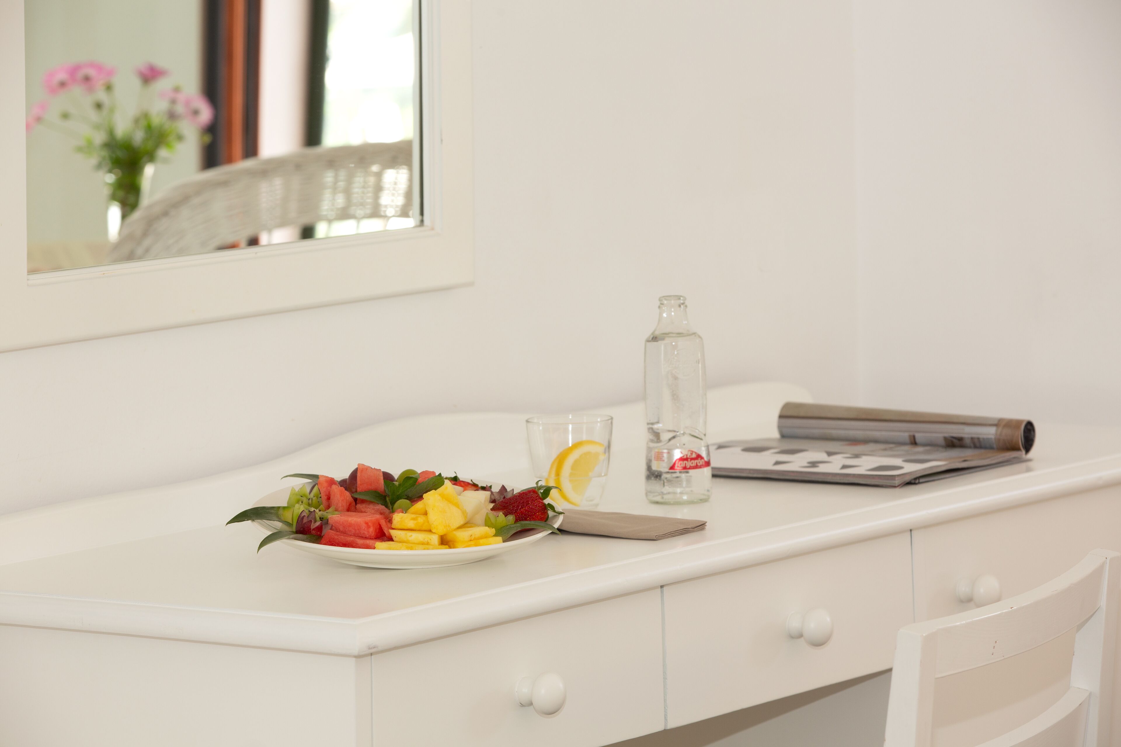Un escritorio blanco con un plato de fruta, un vaso de agua y una botella de agua.